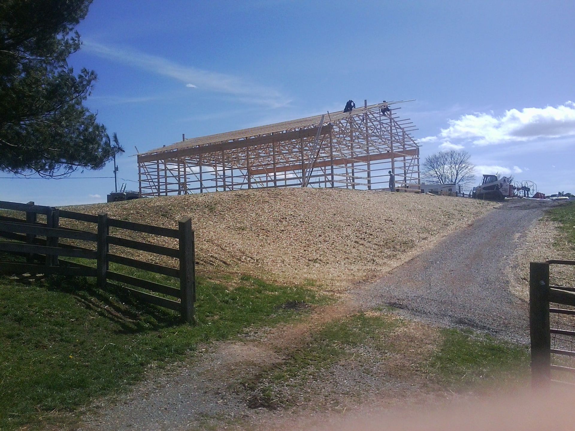 Barn frame under construction on a grassy hill beside a gravel driveway and wooden fence under a blue sky