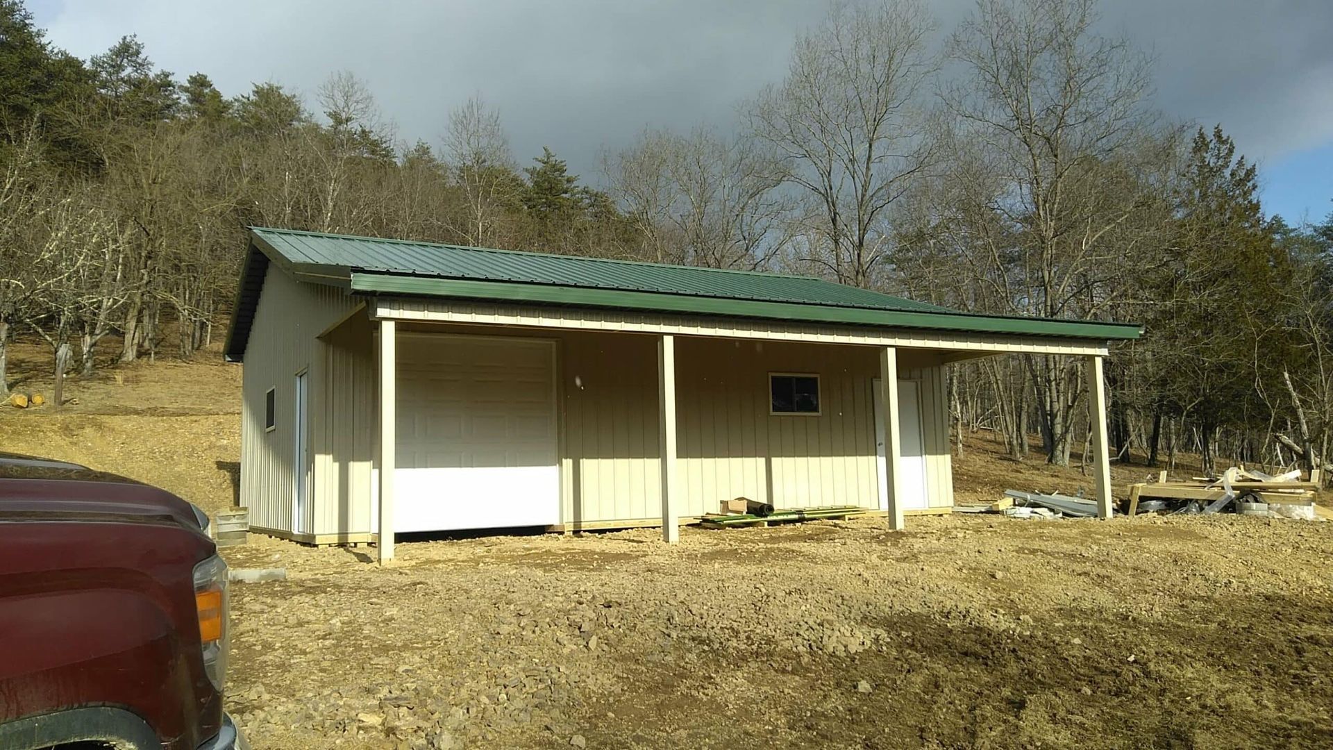 Small beige shed with green roof in a dry wooded clearing, with a red car partly visible at left.