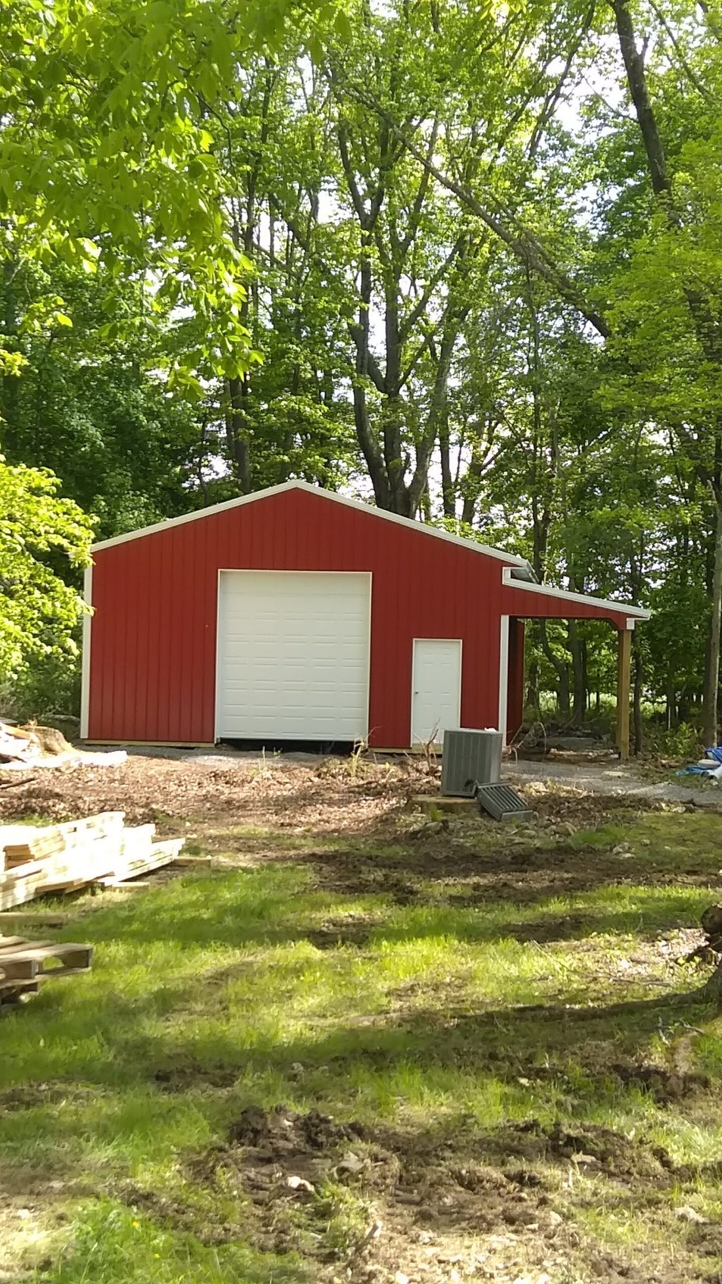 Red shed with white garage door in a wooded yard under trees, with a small covered porch on the right.