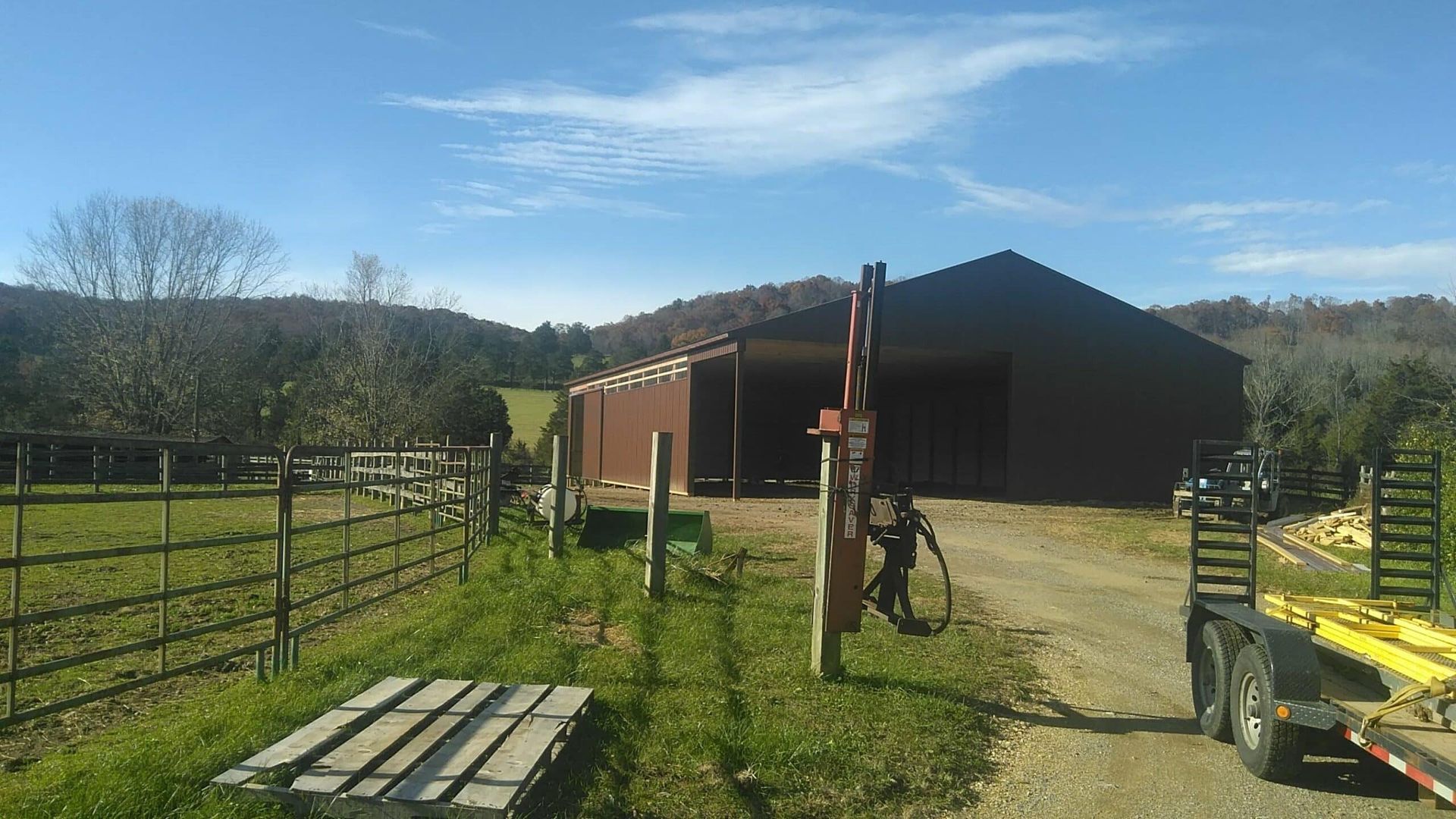 Rural barnyard with a dark barn, fenced paddock, grassy path, and blue sky on a sunny day