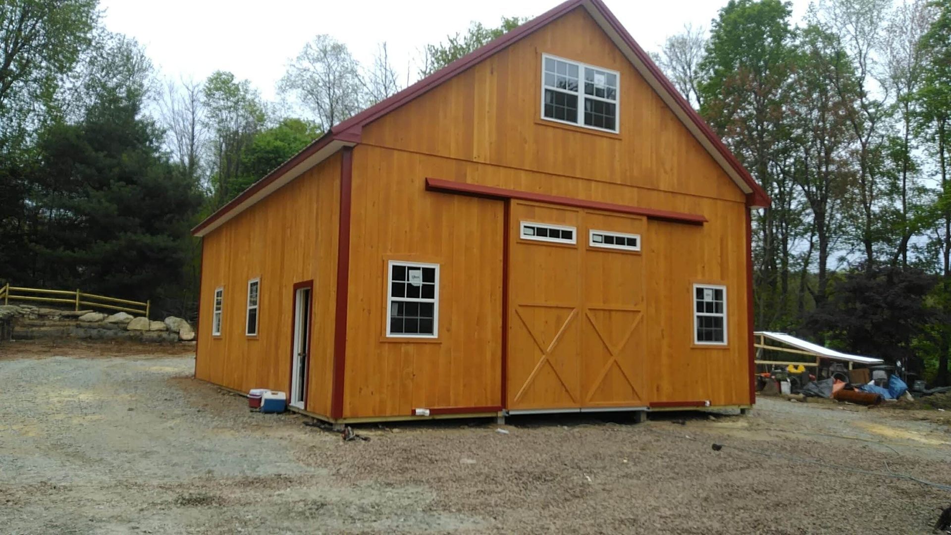 Orange barn-style shed with white-trimmed windows on a gravel lot, surrounded by trees.