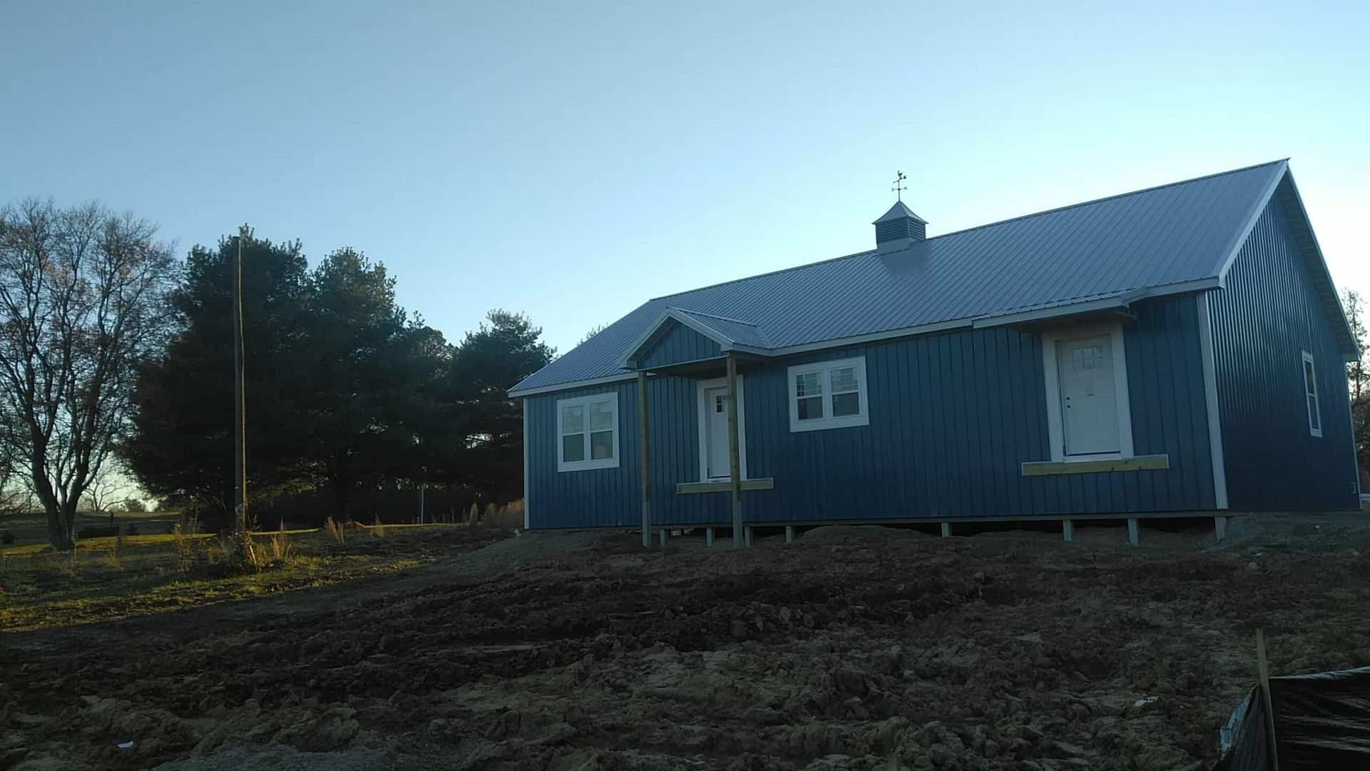 Blue house with white trim in a rural field at dusk, with trees and a dirt foreground.