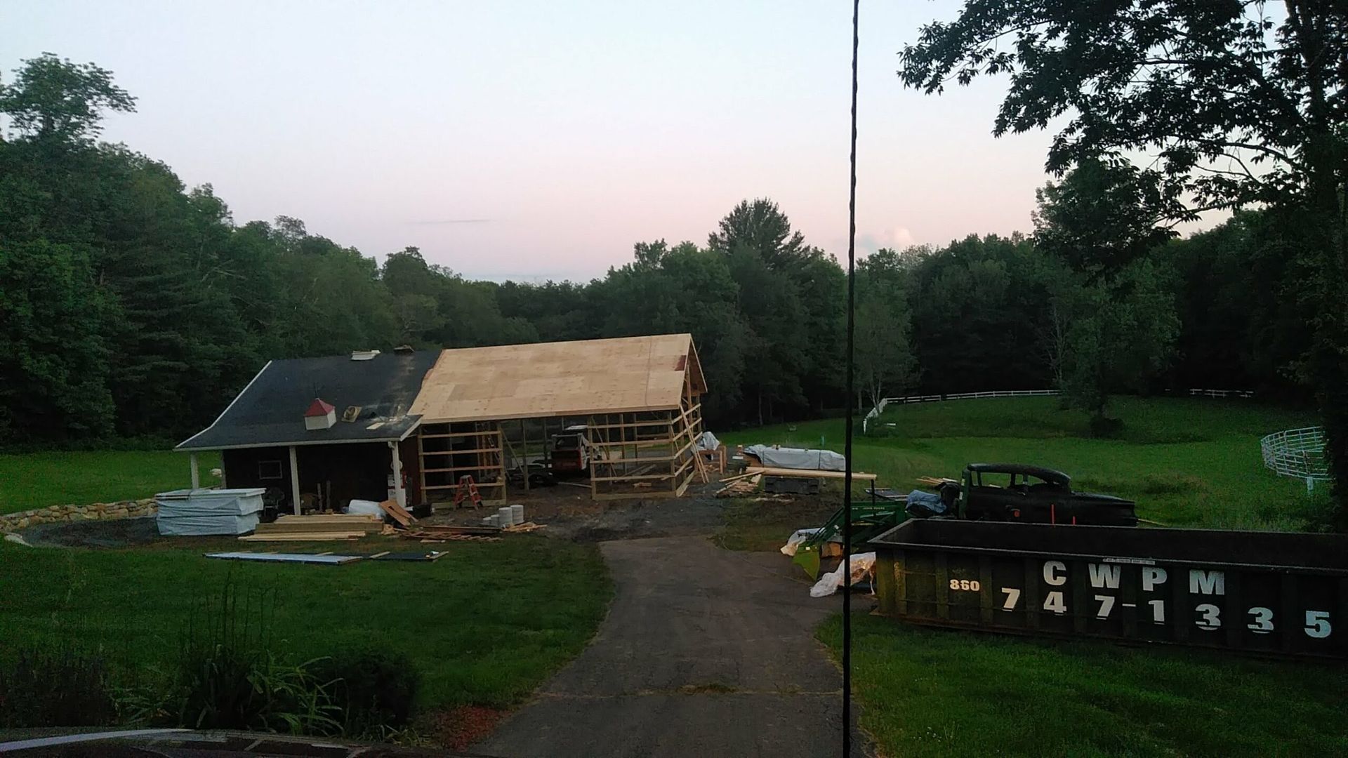 Rural yard with a small wooden shed, stacked firewood, and a driveway at dusk, surrounded by trees.