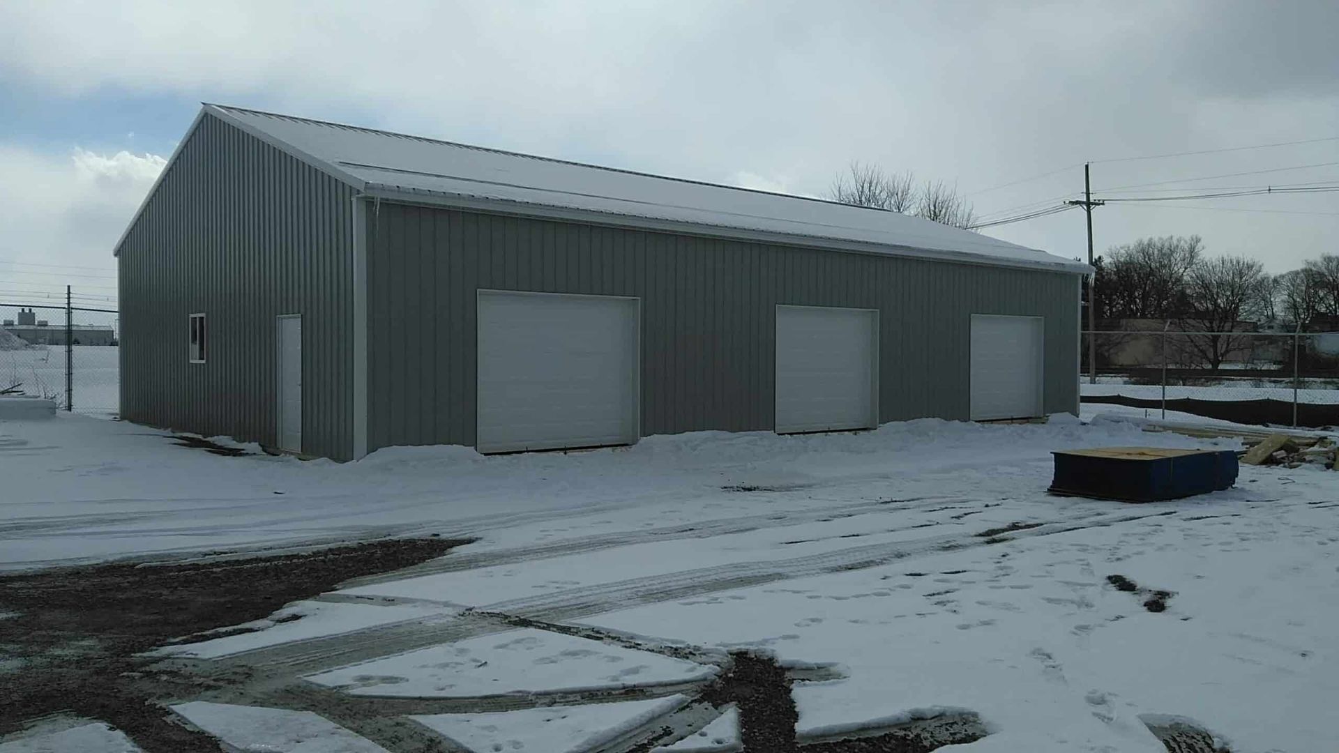 Gray metal warehouse with garage doors in a snowy lot under an overcast sky