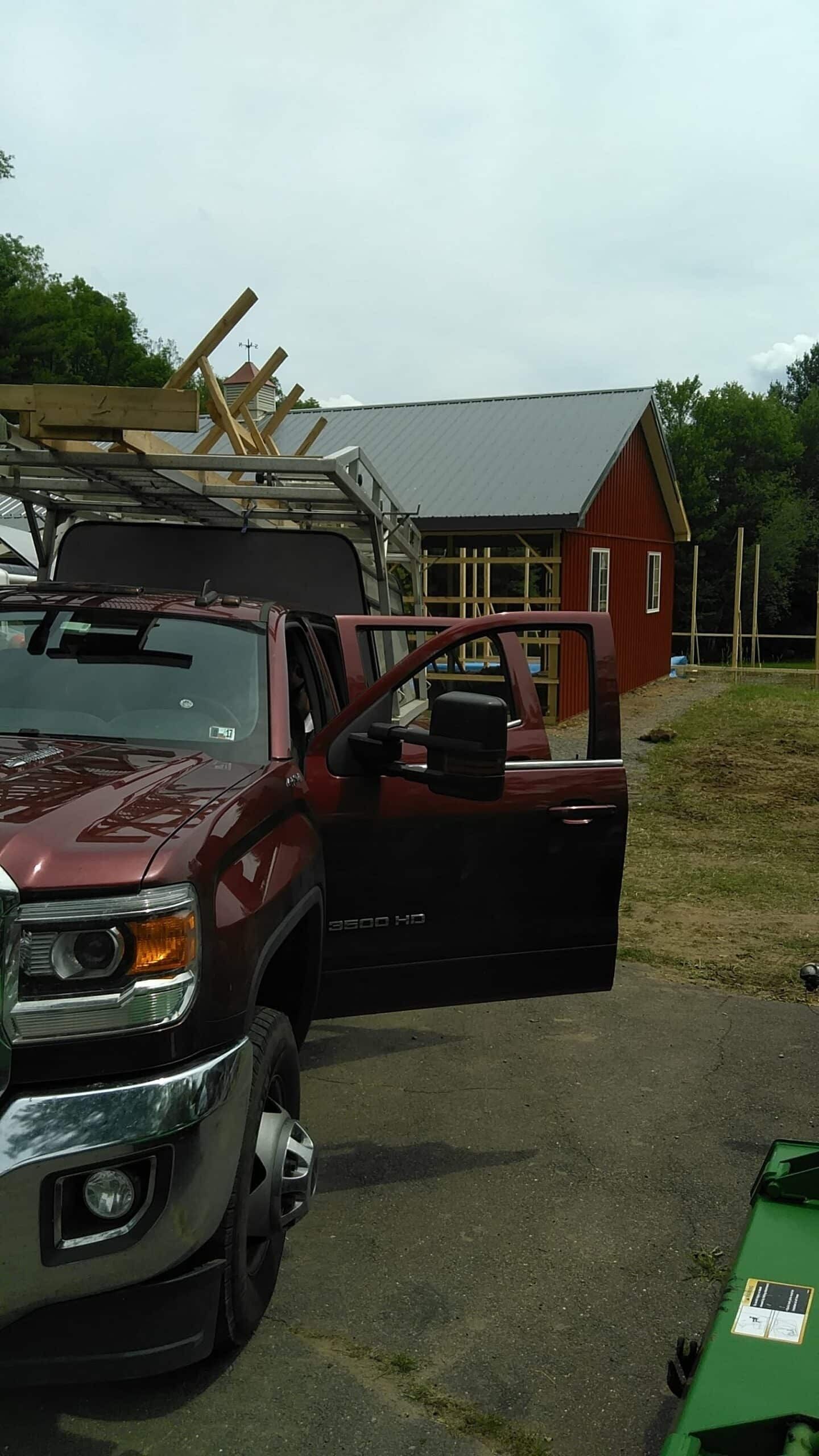 Red truck with an open door and ladder on top parked beside a red building and trees.