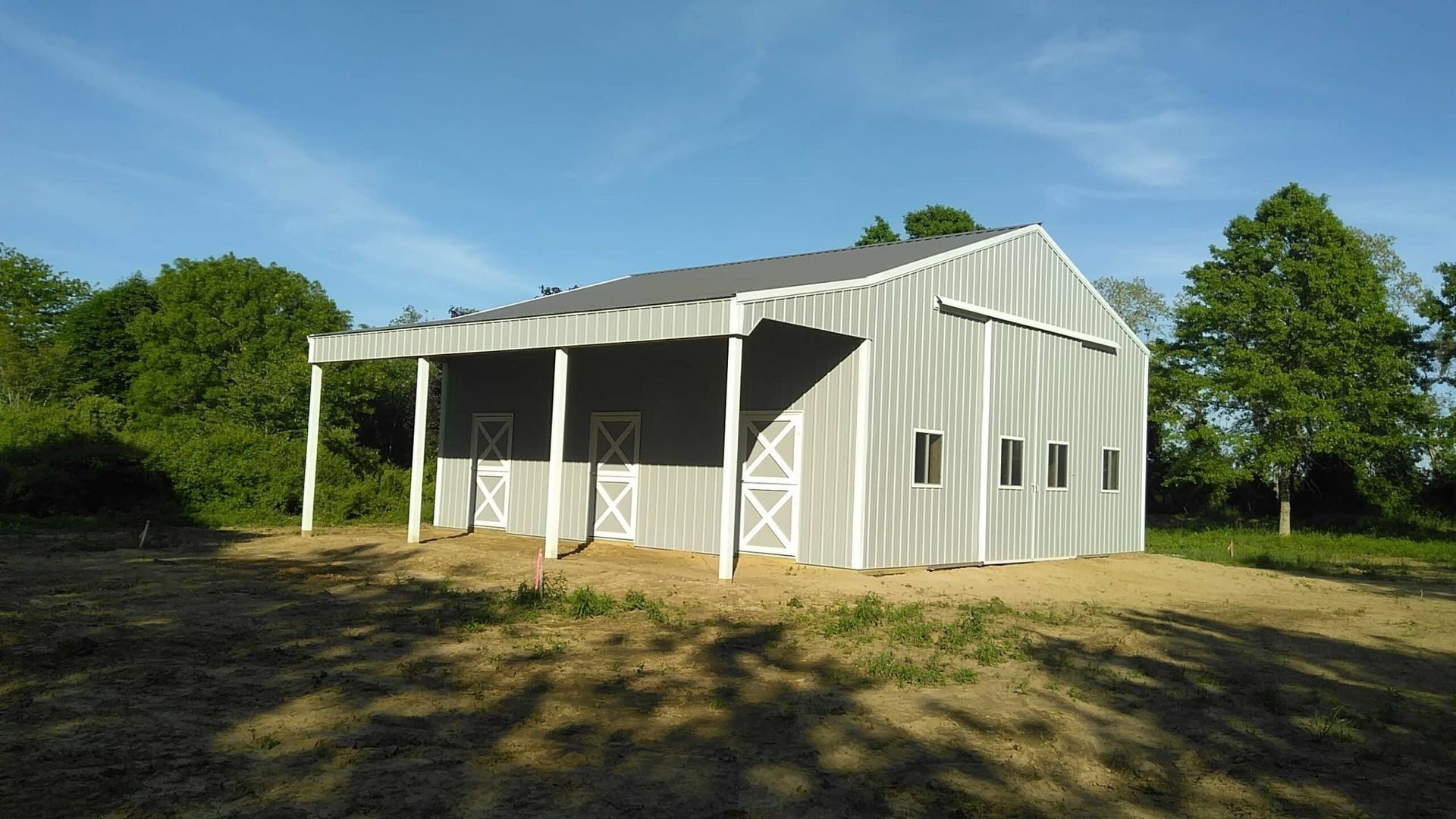 White metal barn with open front and side windows, sitting on a grassy lot beside trees.