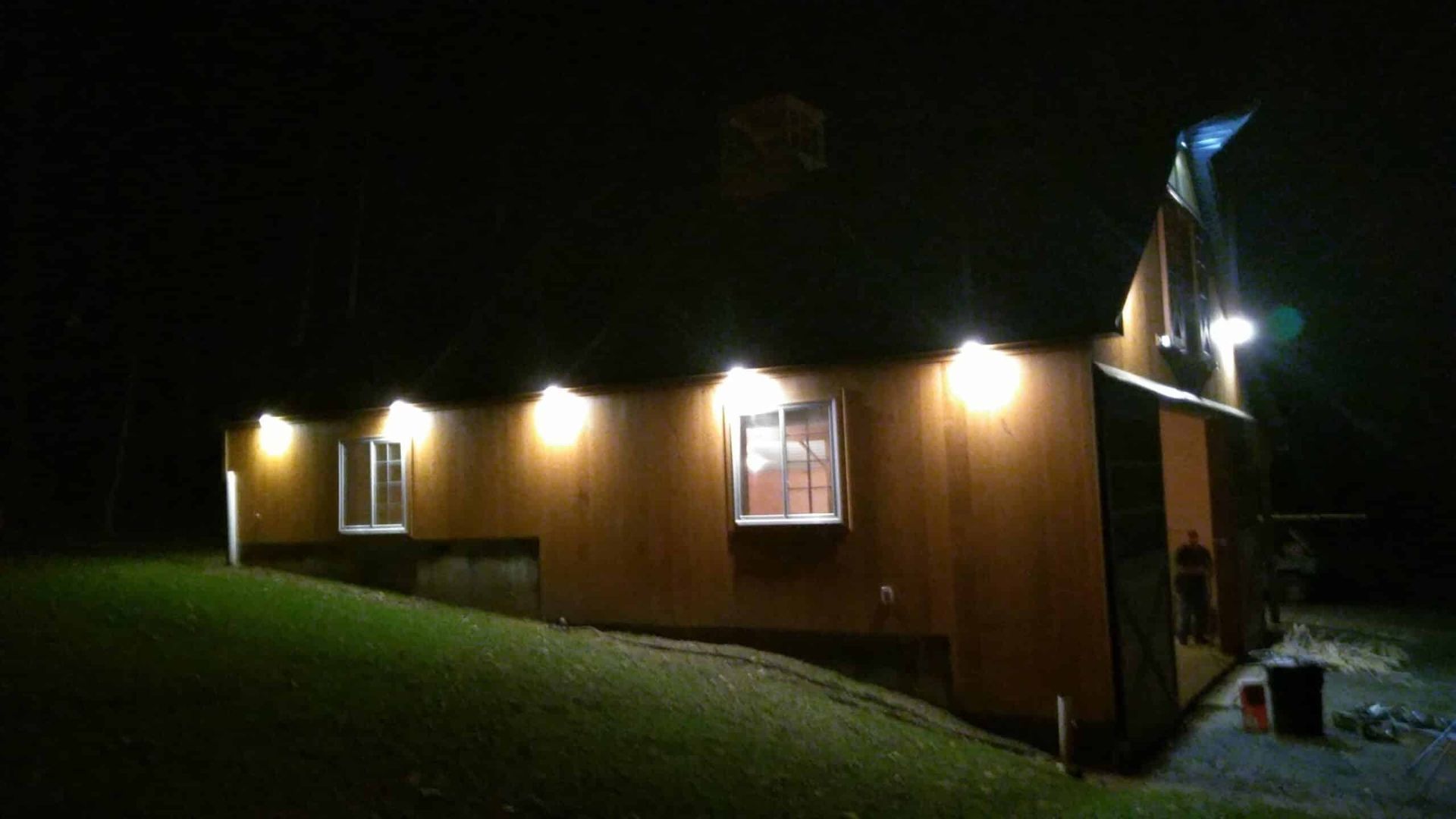 Nighttime view of a lit wooden house on a grassy slope, with lights glowing from the windows.