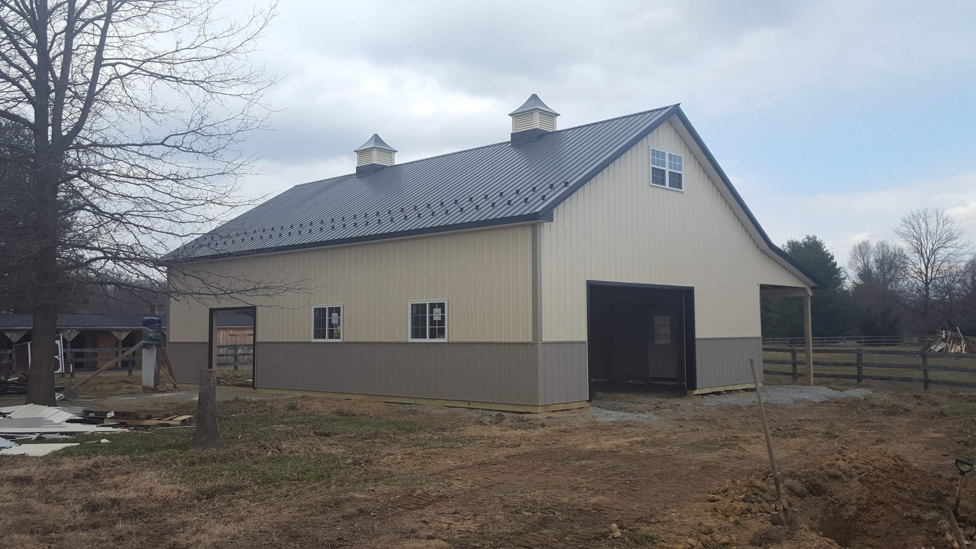 Beige metal barn with white roof and open doorway in a rural yard under cloudy sky