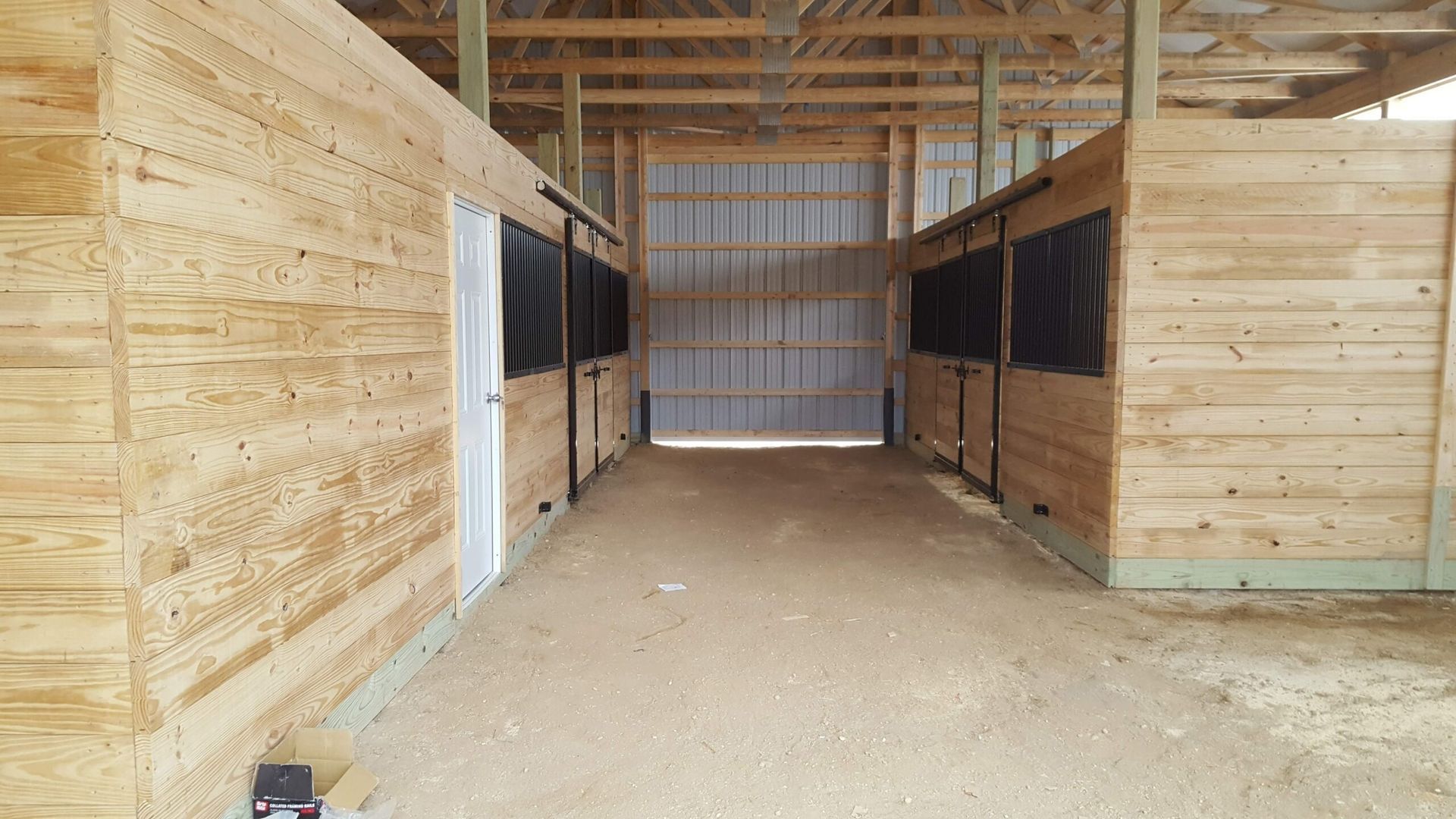 Interior of a barn aisle with wood walls, open stalls, and a dirt floor under exposed rafters.