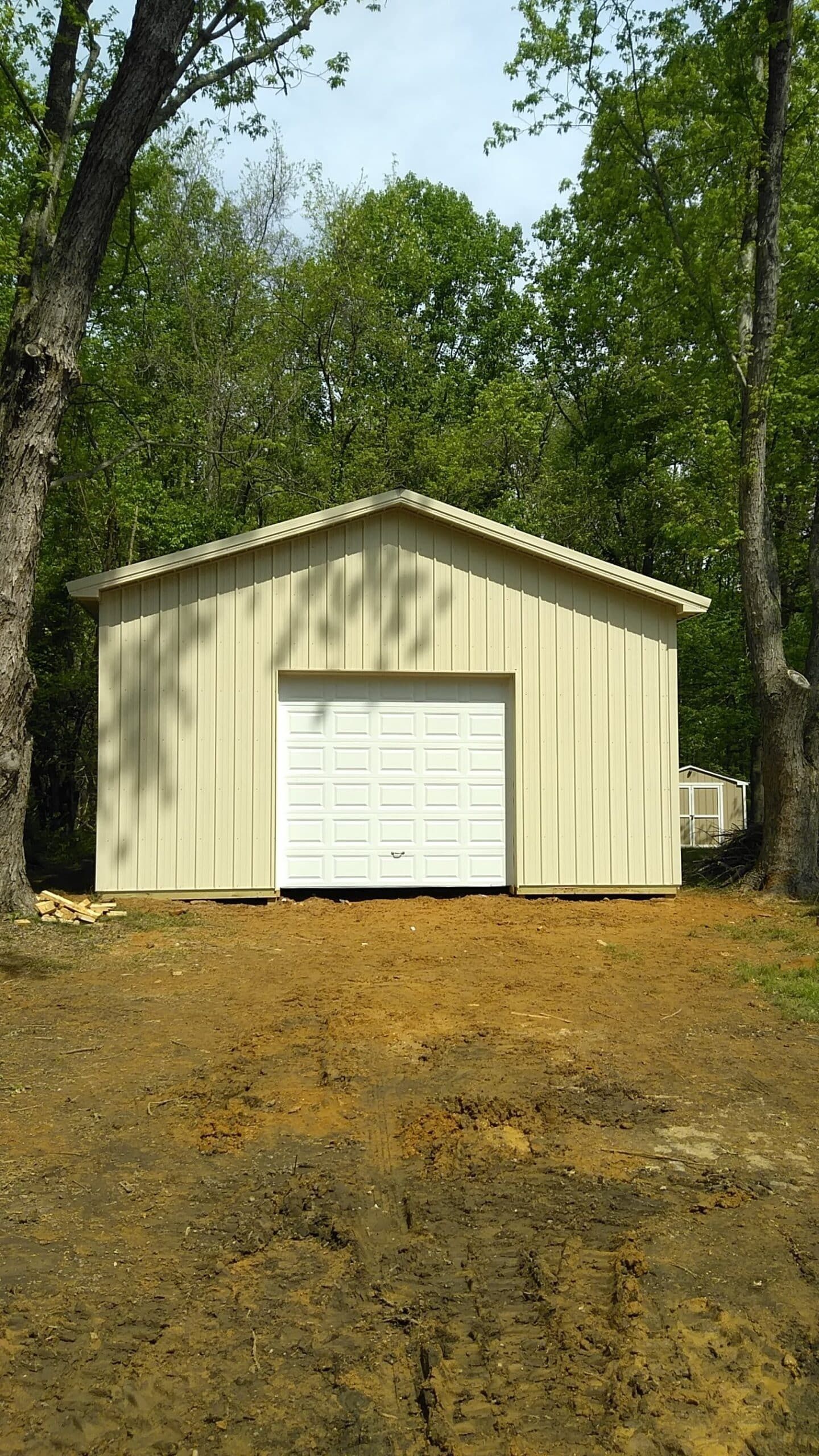 Cream-colored garage with a white roll-up door in a wooded yard, viewed from a dirt driveway.
