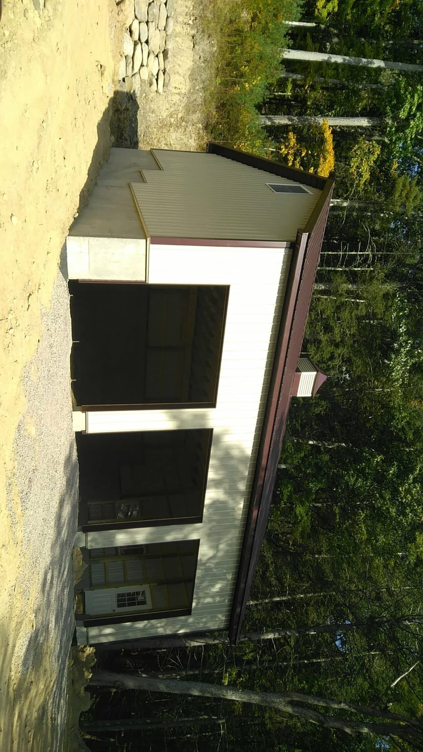 Small white shed with a maroon roof in a wooded yard, viewed from the side.