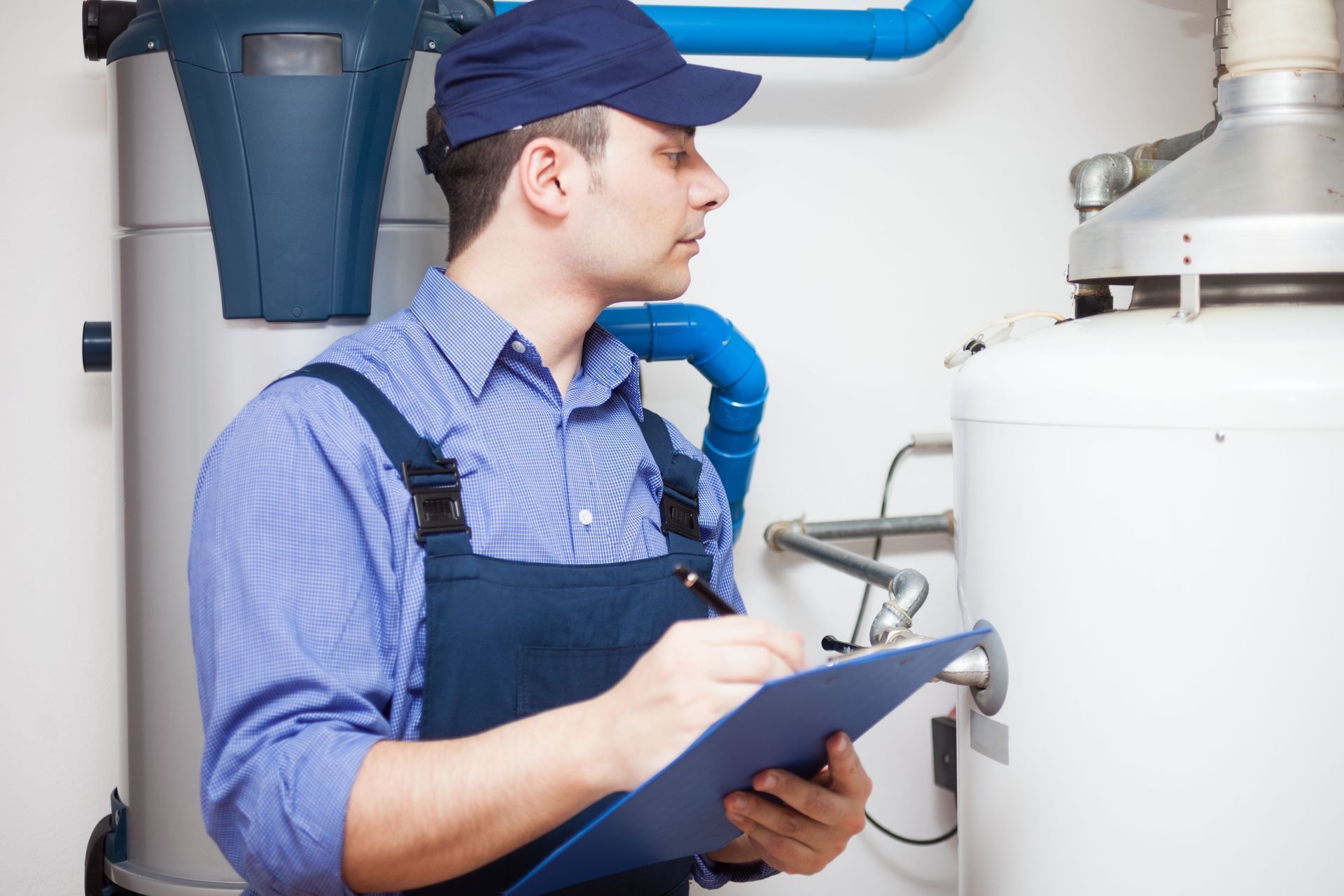 Plumber in blue uniform, examining a water heater and taking notes on a clipboard.