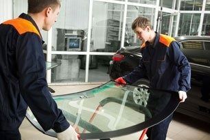 Two mechanics in blue jumpsuits holding a car windshield in a repair shop.