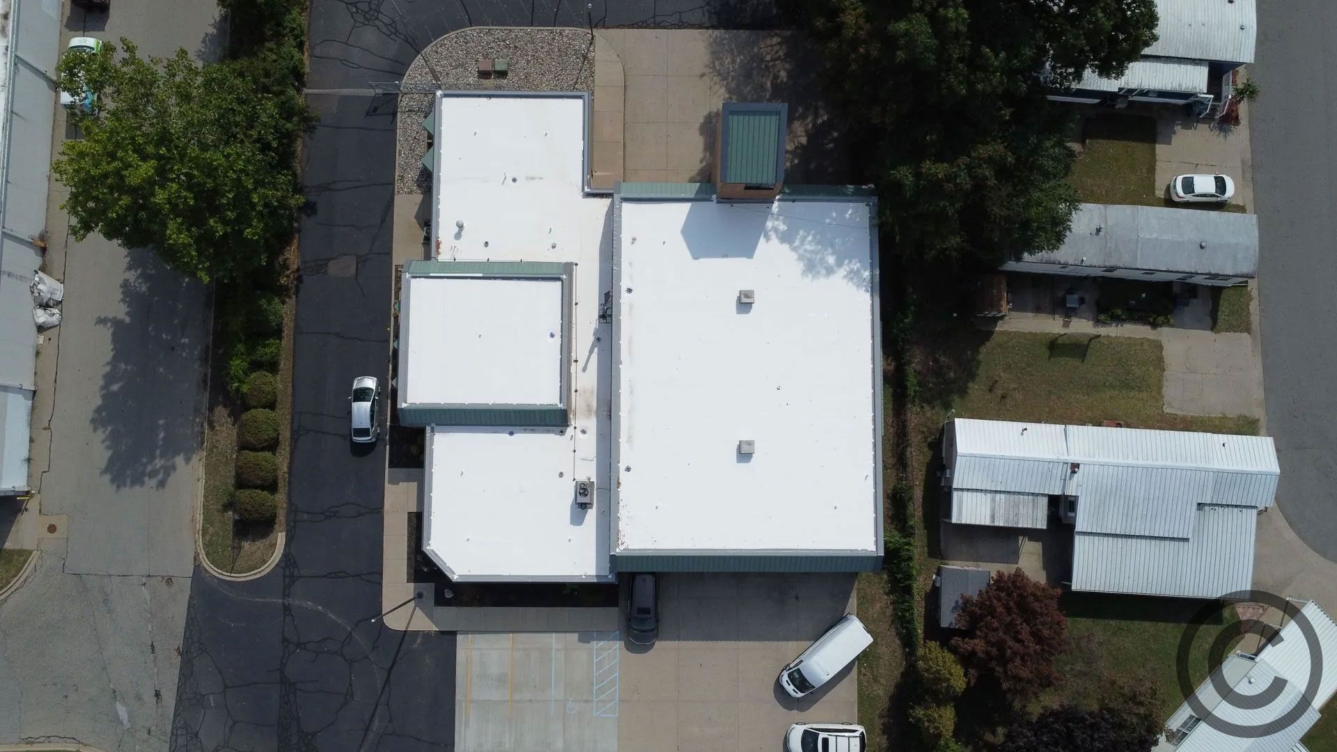Aerial view of a commercial building with a white roof, surrounded by parking and trees.
