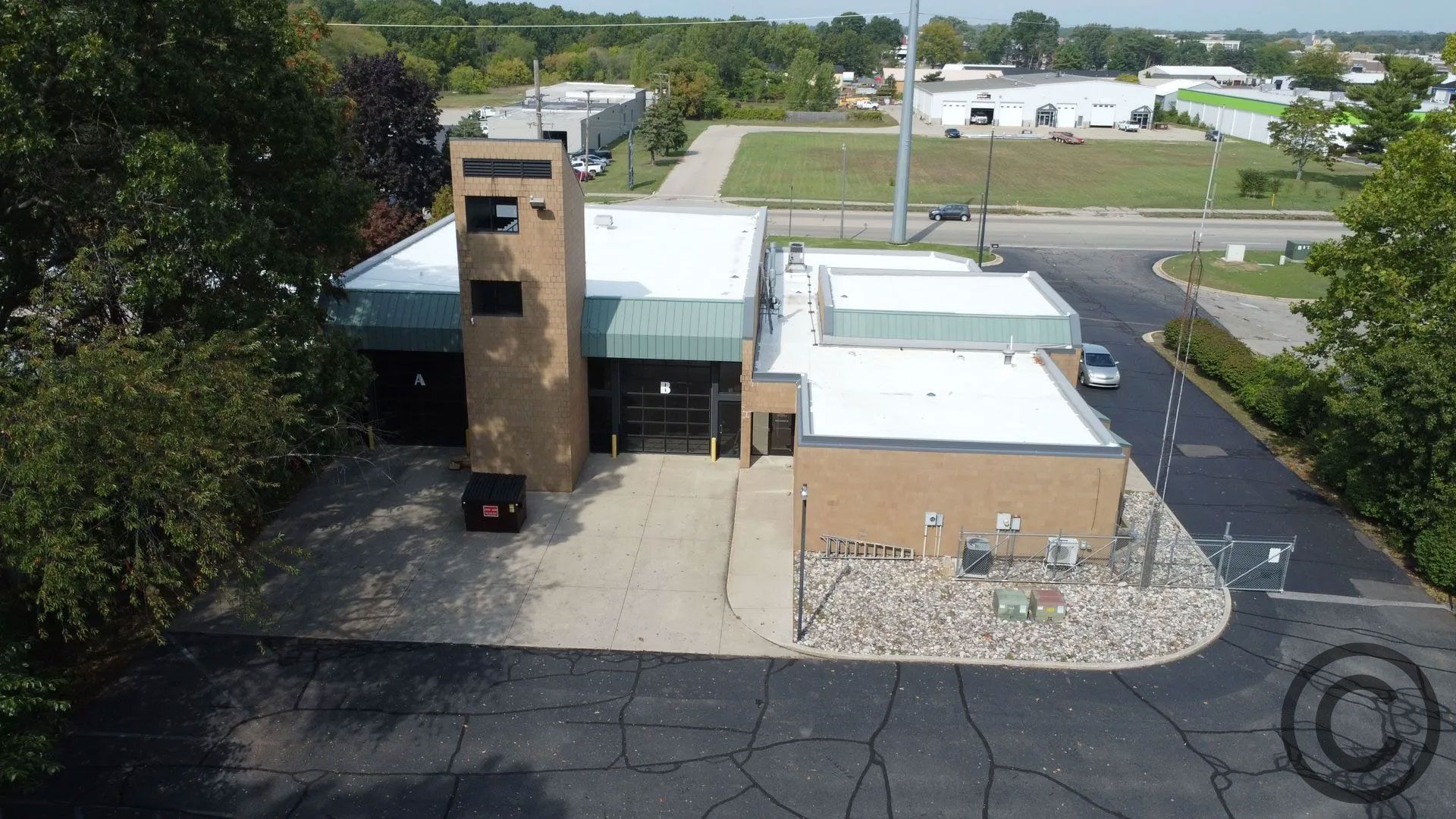 Fire station with tan brick exterior, tall tower, and black garage doors; surrounded by parking lot and trees.