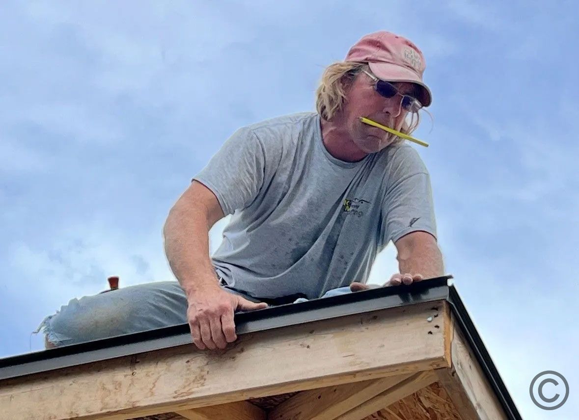 Man on a roof wearing a pink cap, chewing a pencil, working on the edge, against a cloudy sky.