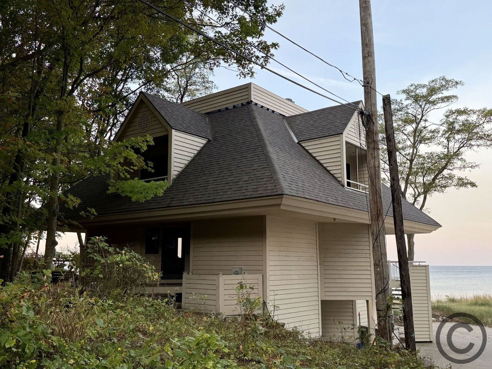 Two-story, white house with dark roof near a shoreline. Green trees and shrubs surround.