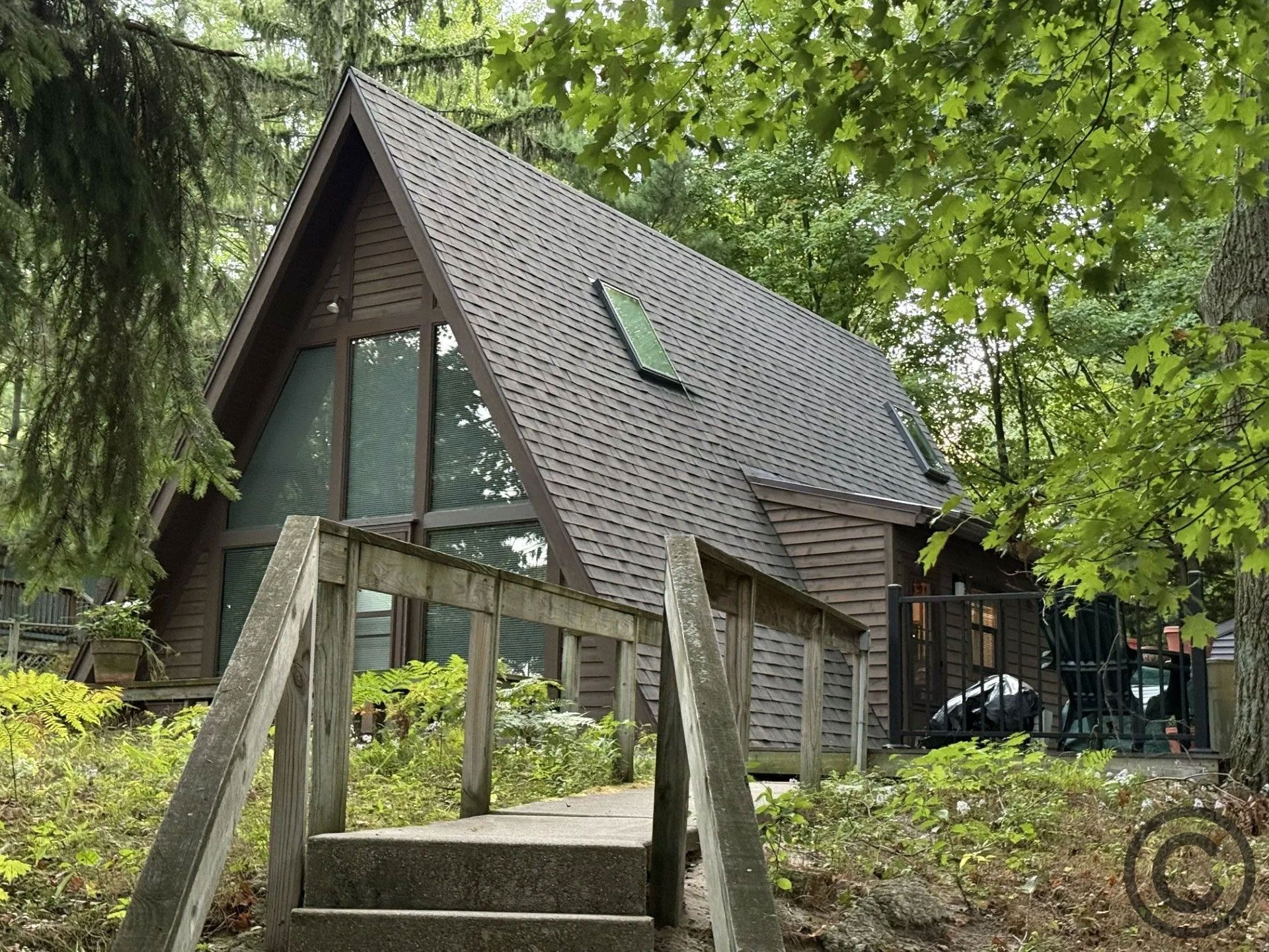 A-frame cabin nestled in trees, reached by a wooden bridge. Brown siding and roof, large windows.
