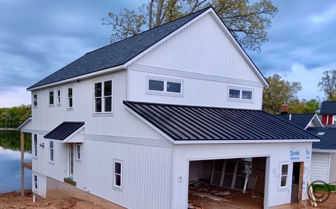 White house with black roof and garage on water's edge under cloudy sky.