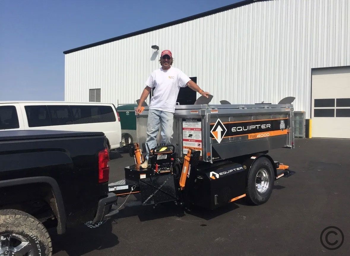 Man stands in a dump trailer hitched to a truck outside a building on a sunny day.