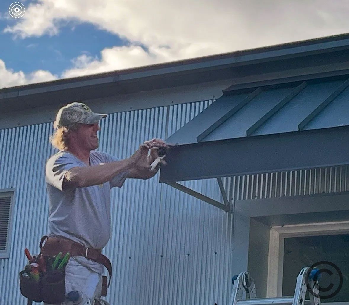 Man on ladder installing a metal awning on a corrugated metal building. Cloudy sky.