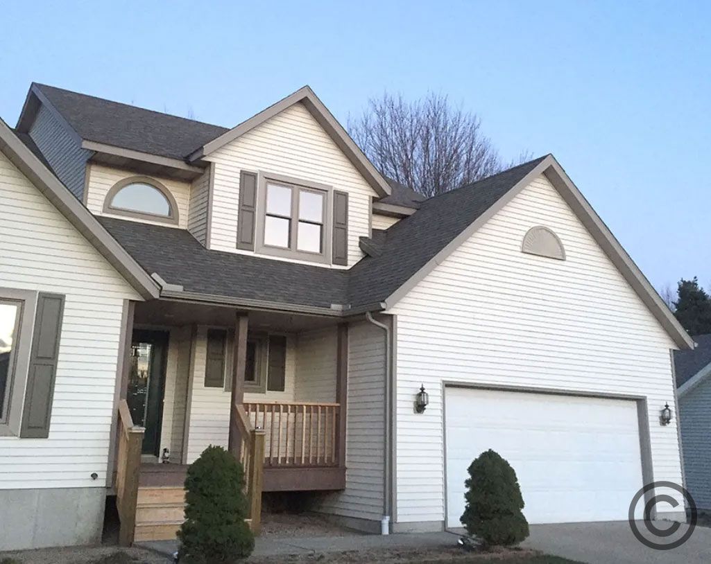 Two-story house with white siding, brown roof, and garage door; small front porch and shrubs in front.