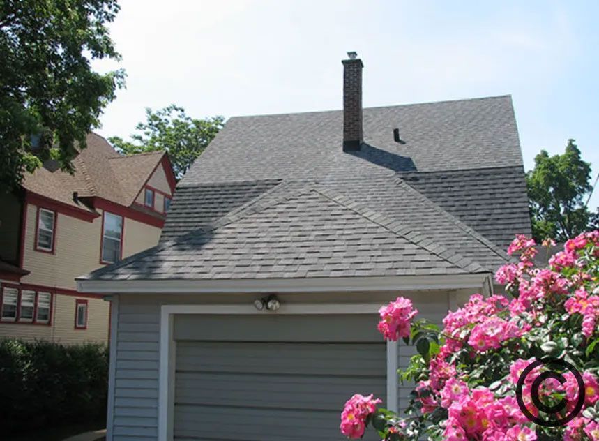 Garage with a gray roof, chimney, and blue siding, with pink roses in the foreground and a house in the background.