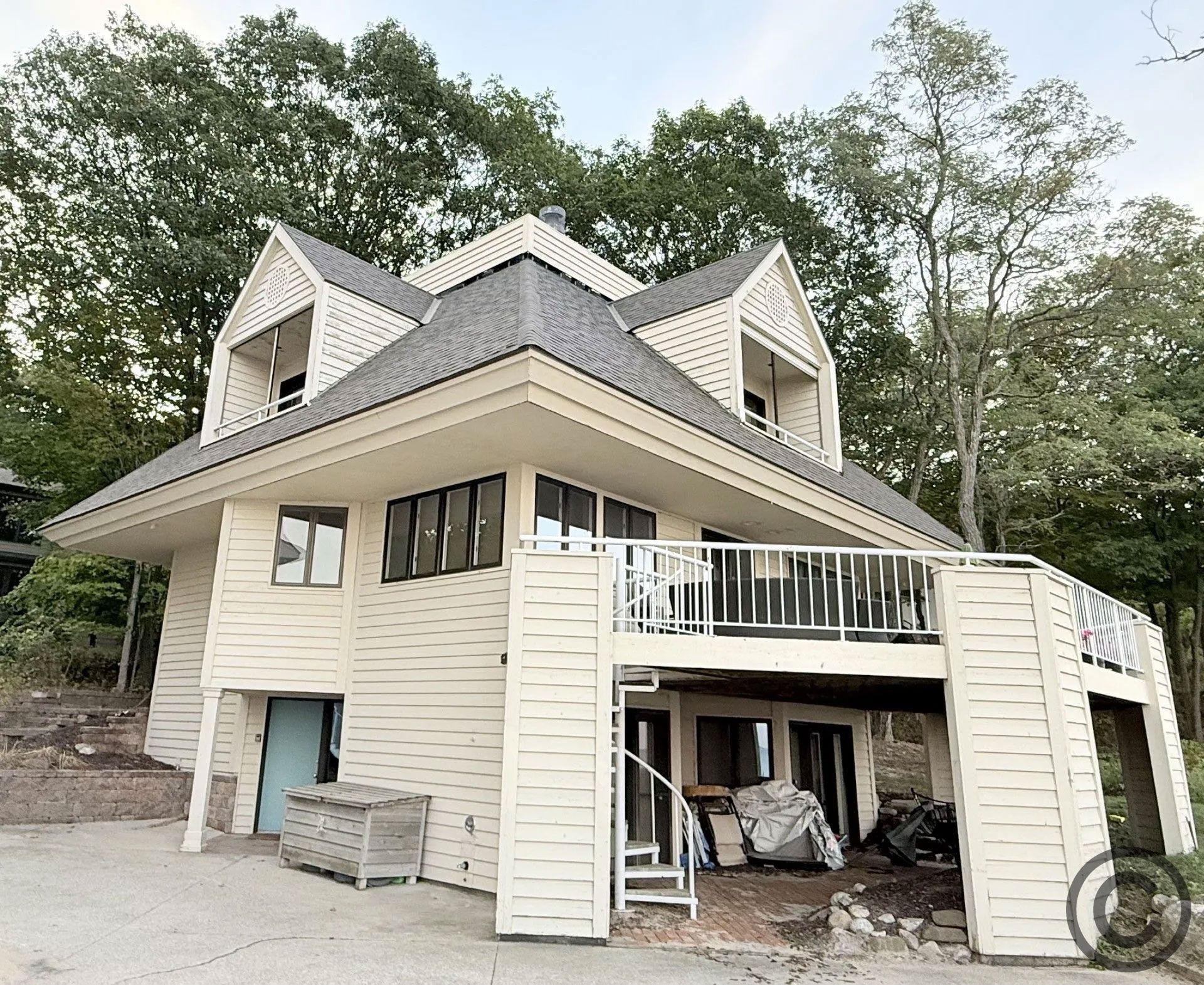 Two-story house with a balcony, siding and an A-frame roof, nestled amongst trees.