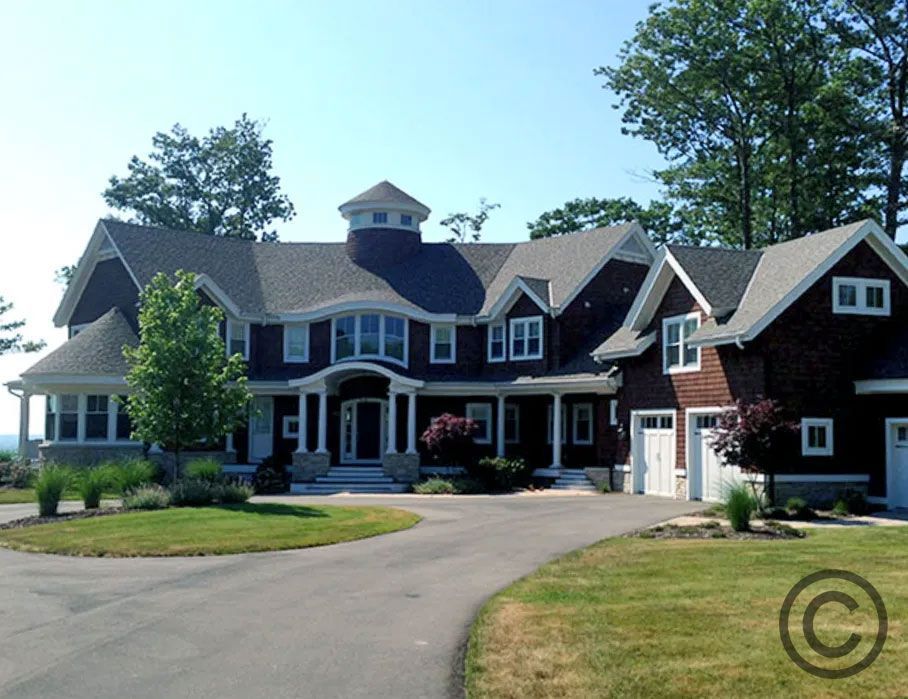 Large, two-story brown house with white trim, circular driveway, green lawn, and blue sky.