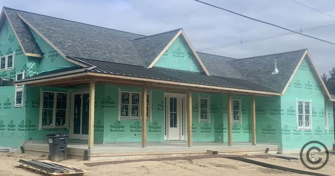 House under construction, green wrap, gray roof, porch, cloudy sky, wood columns.