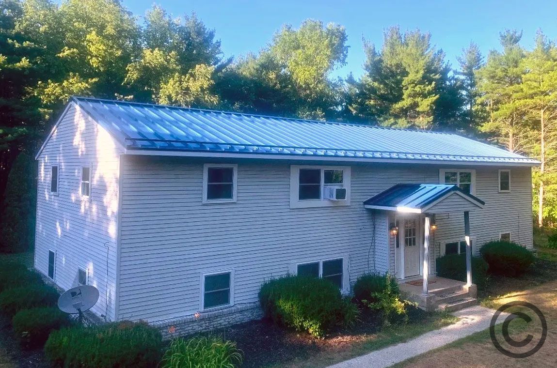 Two-story house with blue metal roof, white siding, and surrounding trees.