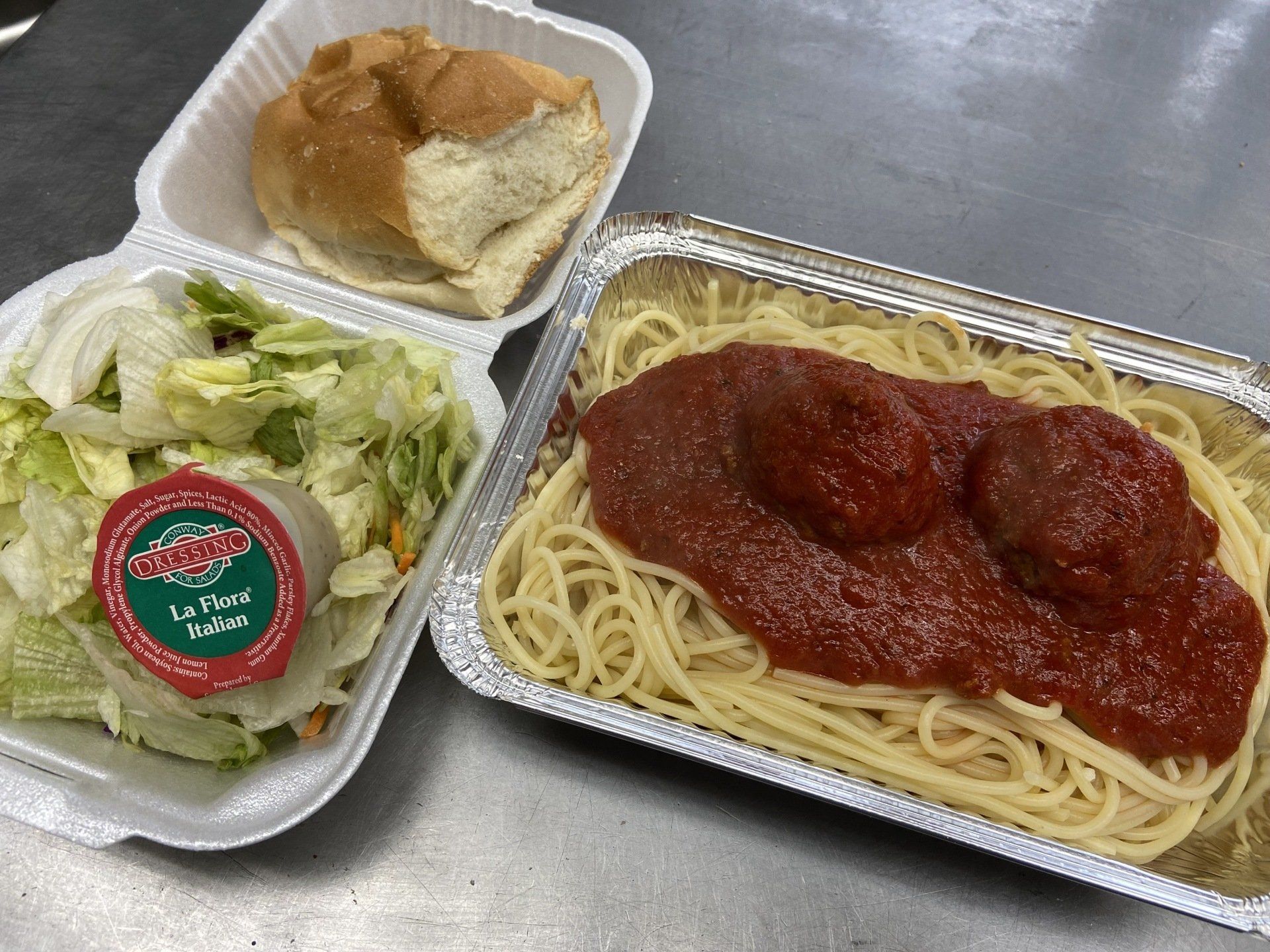 Spaghetti with meatballs, salad, and a roll in take-out containers on a metal surface.