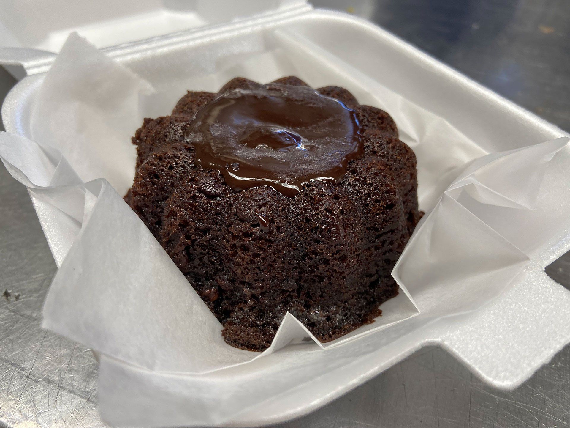 Chocolate cake in a white foam container, with chocolate topping, on white tissue.