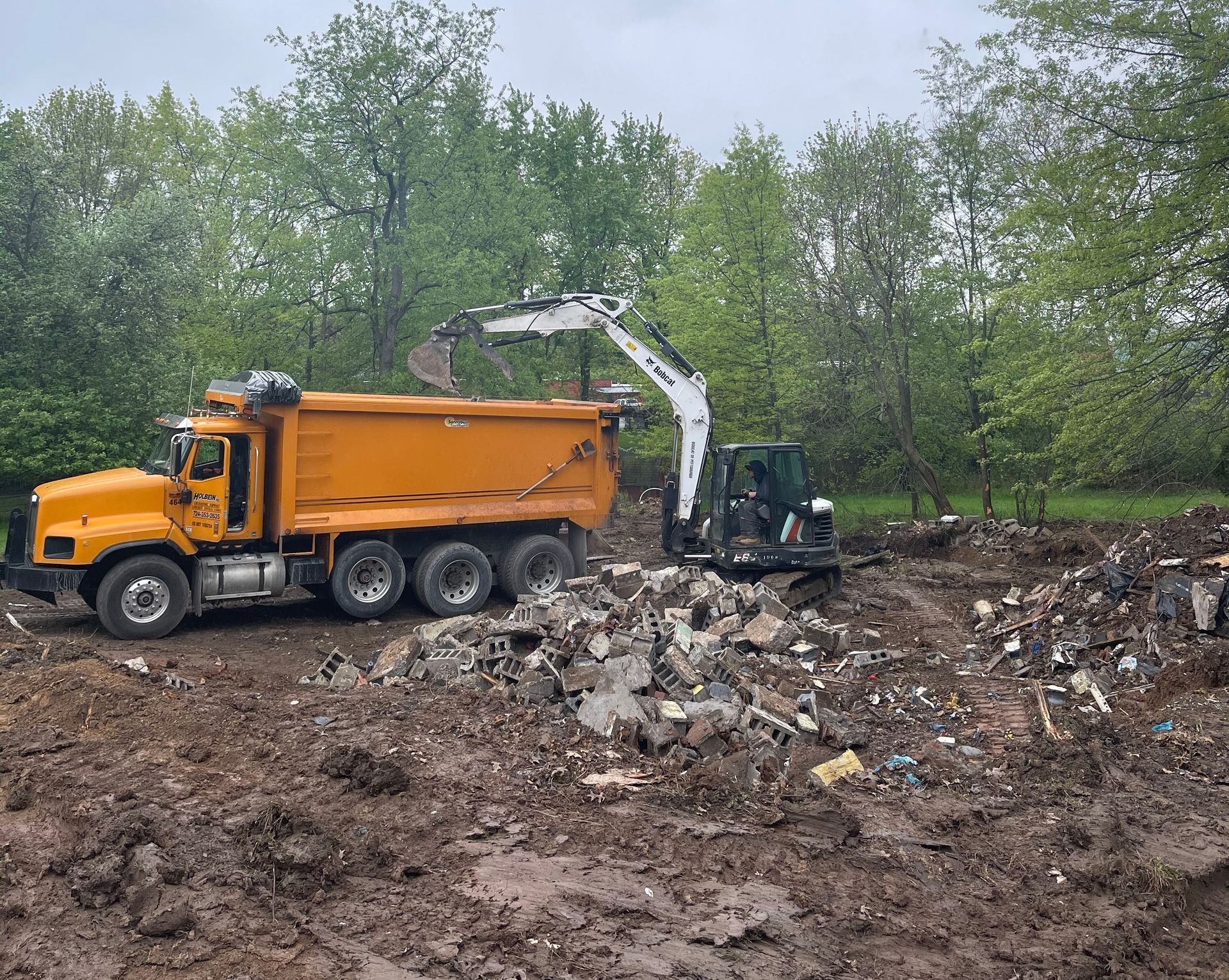Yellow dump truck and excavator loading debris in a muddy field with trees in the background.