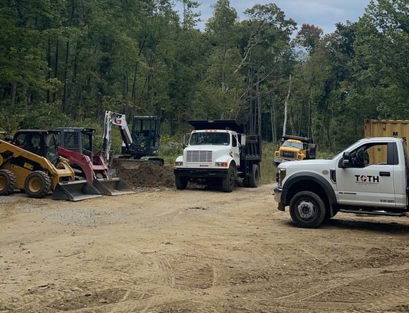 Construction site with heavy machinery, including a dump truck and excavator, against a wooded background.