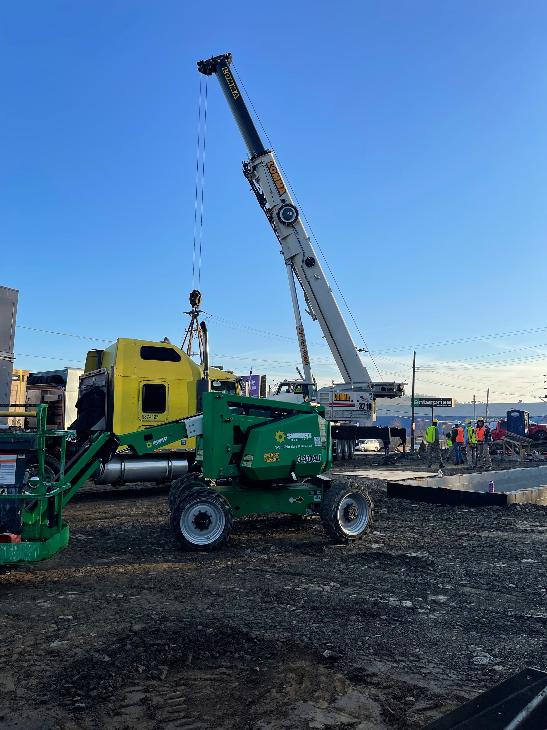 A crane lifting a large object near a yellow truck and a green lift; construction site.