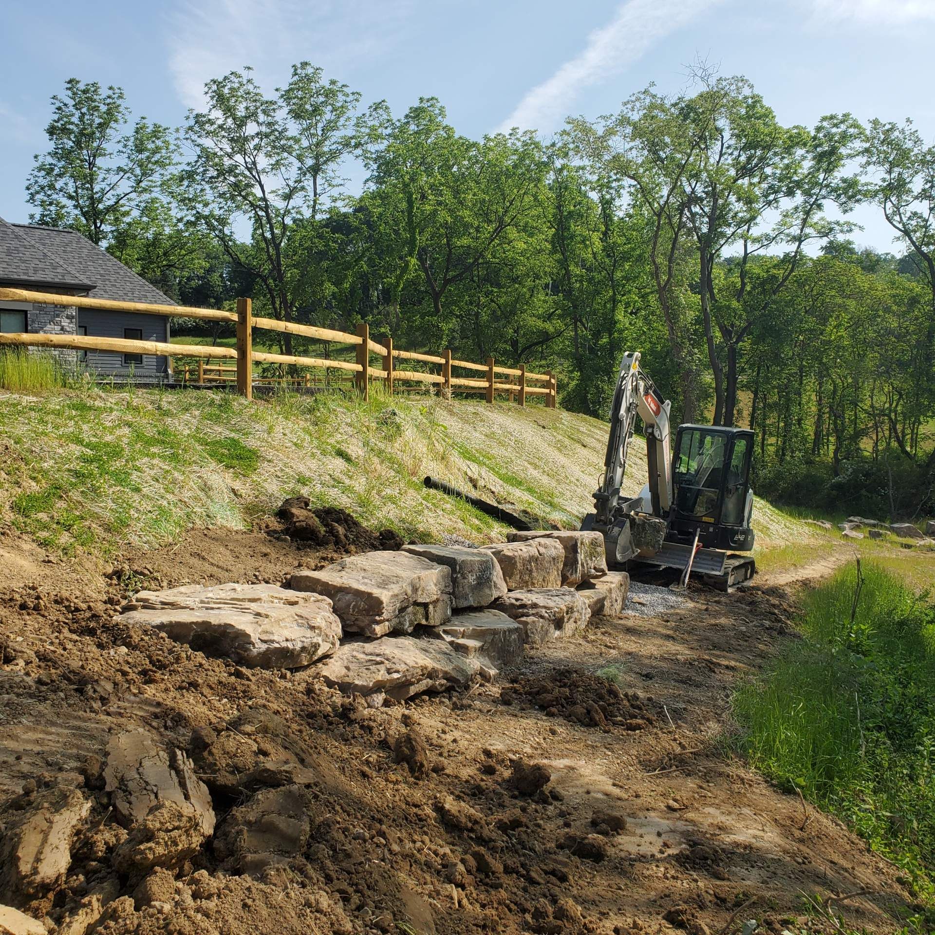 Excavator placing large rocks to build a retaining wall near a building and wooden fence, with green trees.