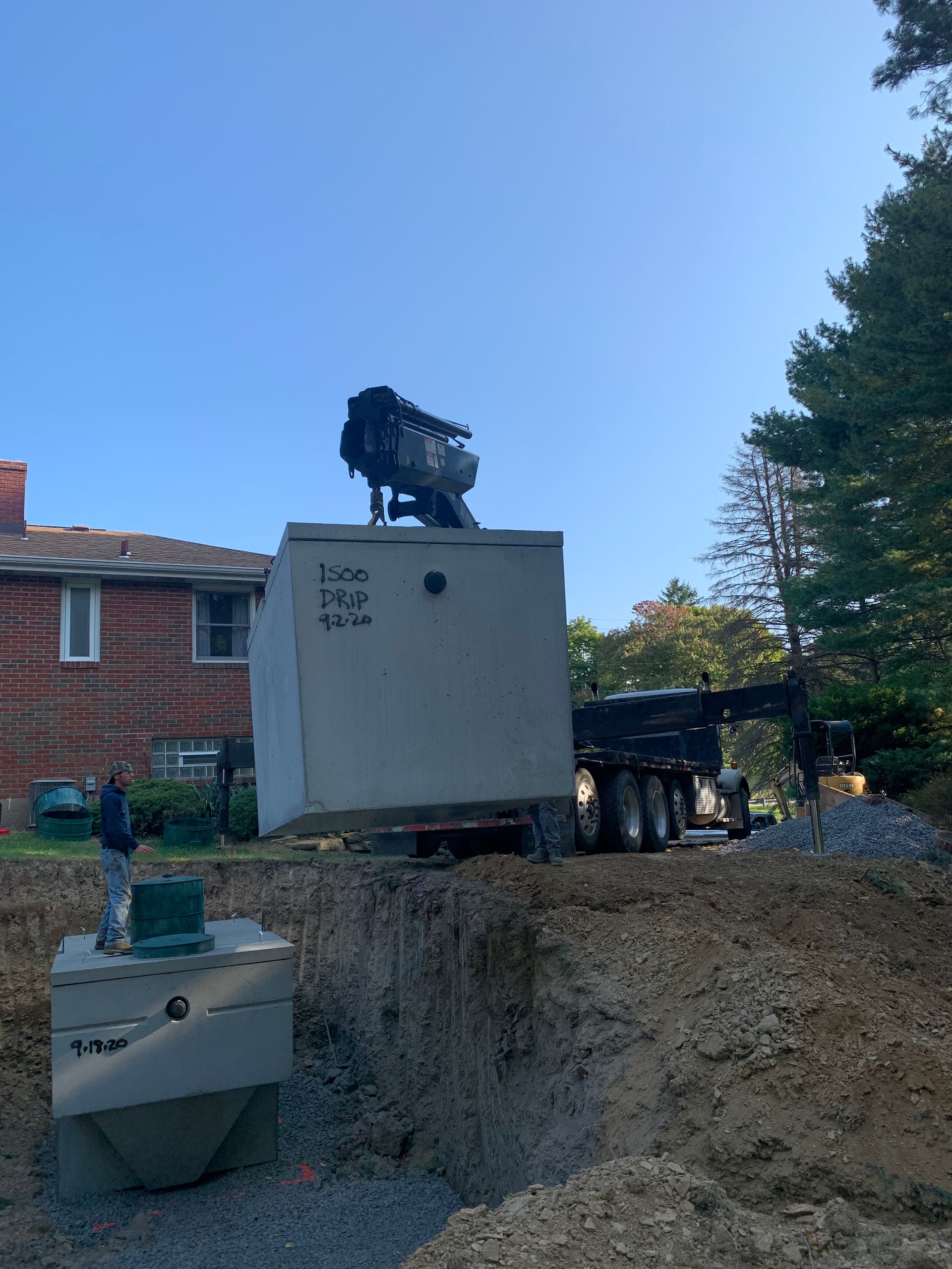 Crane lifting a large concrete septic tank near a house; worker watching. Sunny day.