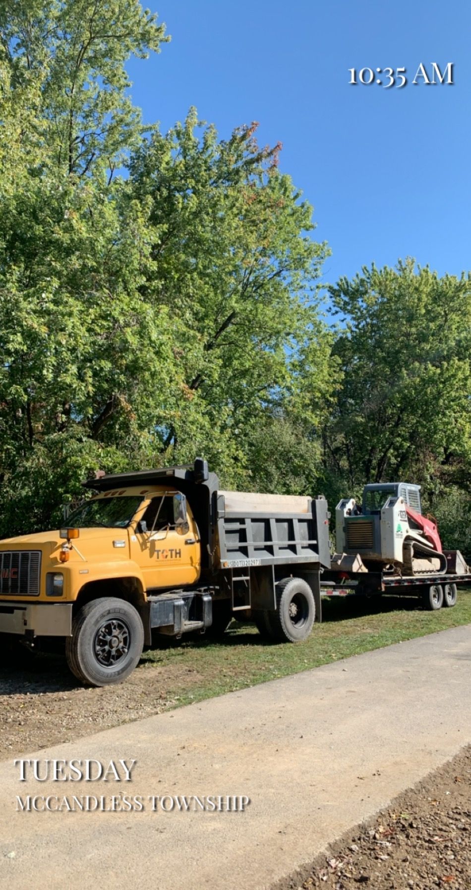 Yellow dump truck with a trailer carrying a skid steer on a road near trees. Sunny day.