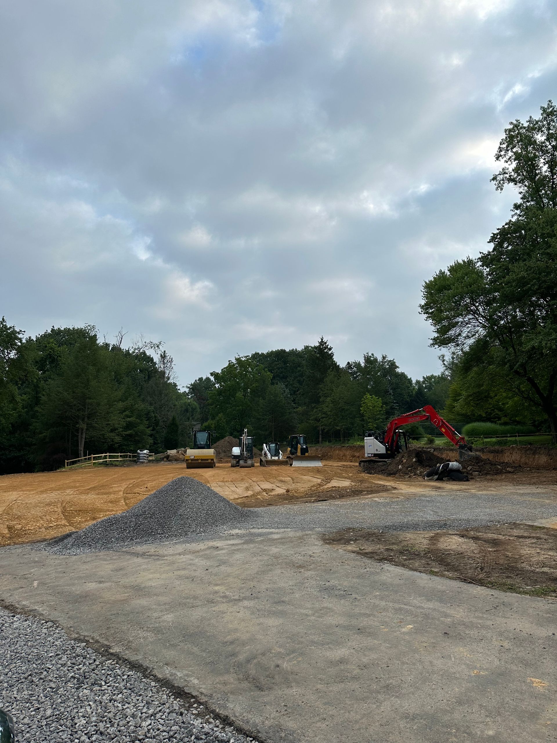 Construction site with gravel road, pile of rocks, excavator, cleared land, and trees under cloudy sky.