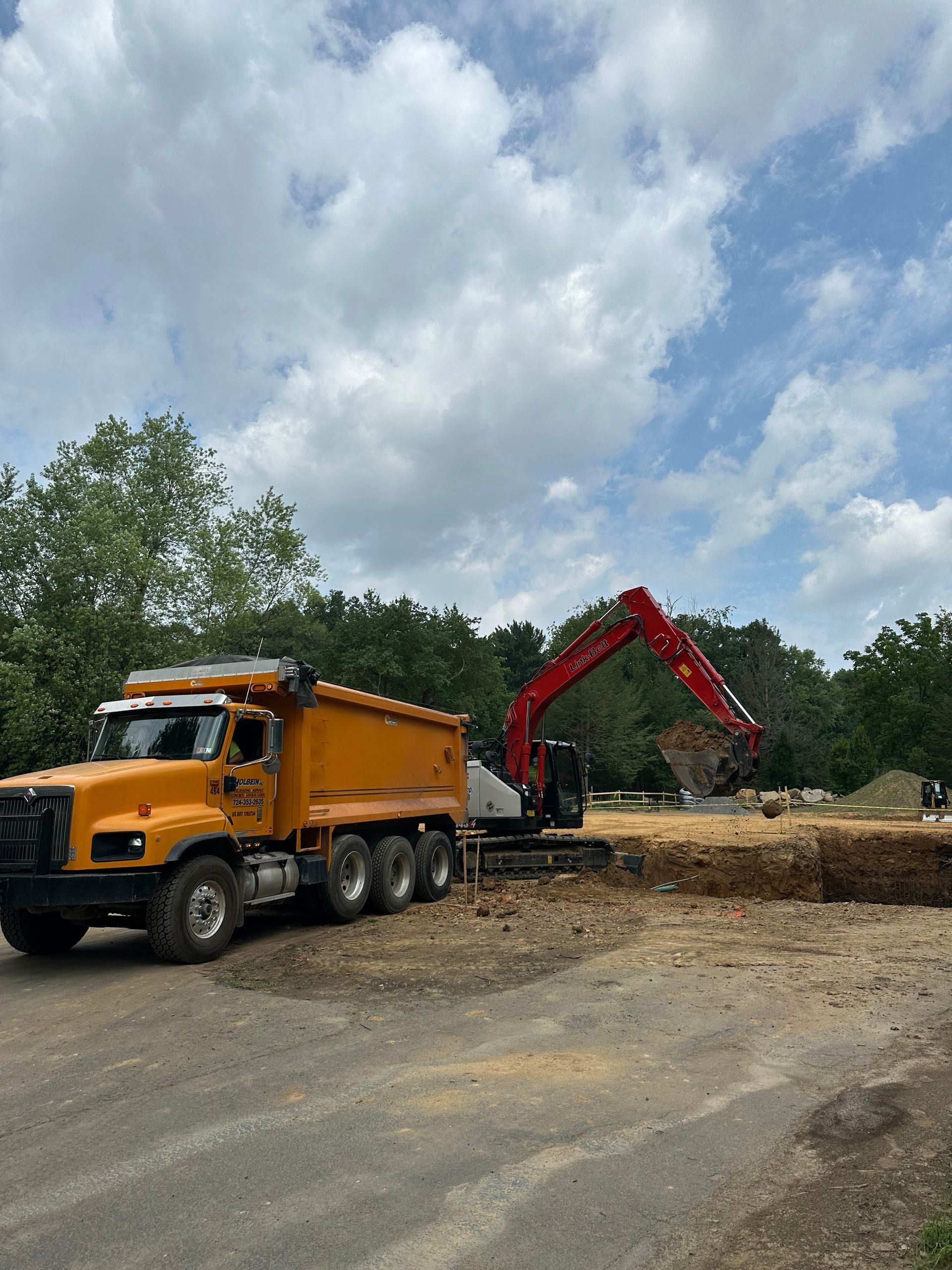 Yellow dump truck next to a red excavator loading dirt under a cloudy sky.