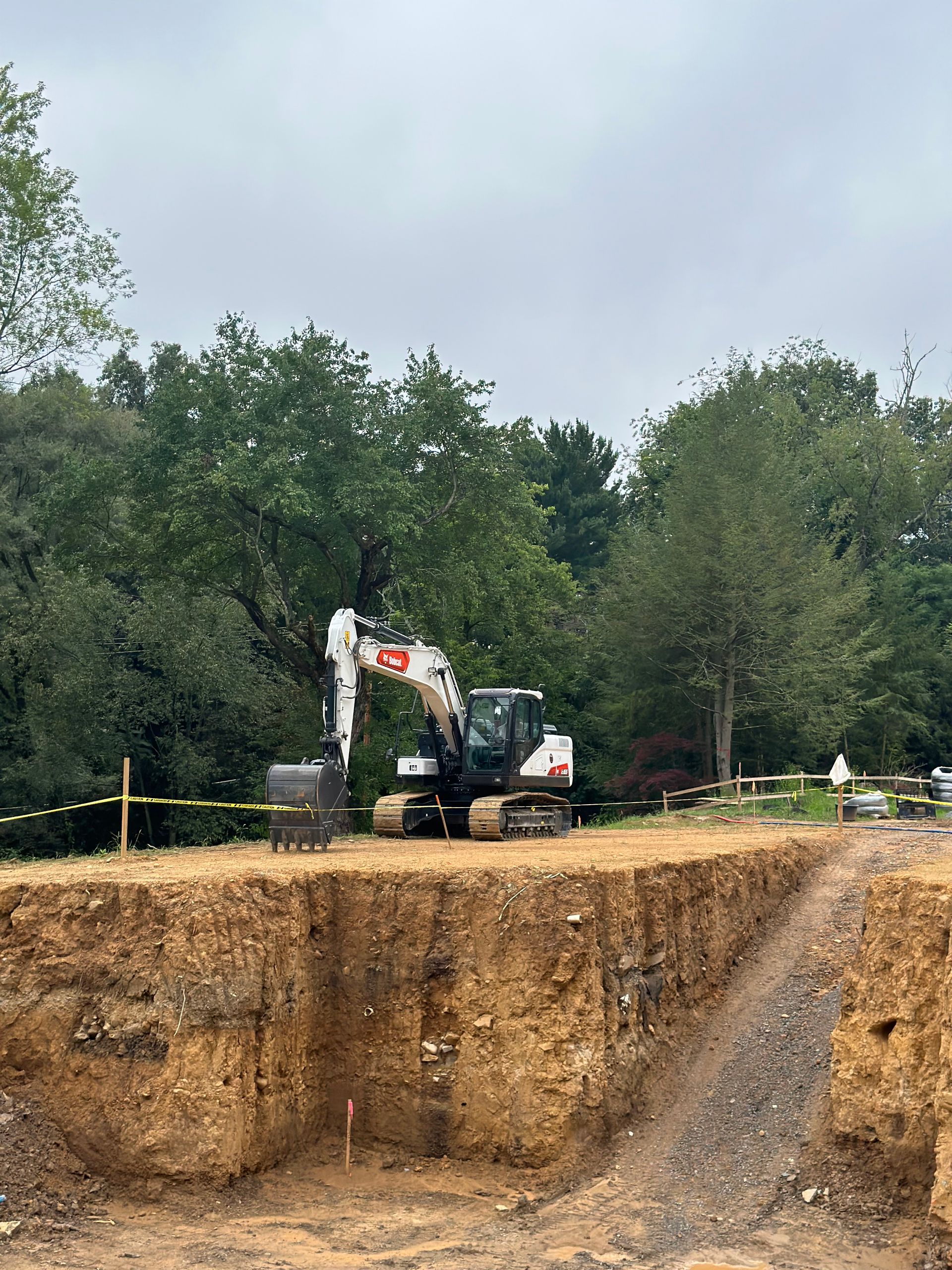 An excavator sits in a large pit at a construction site near trees on an overcast day.