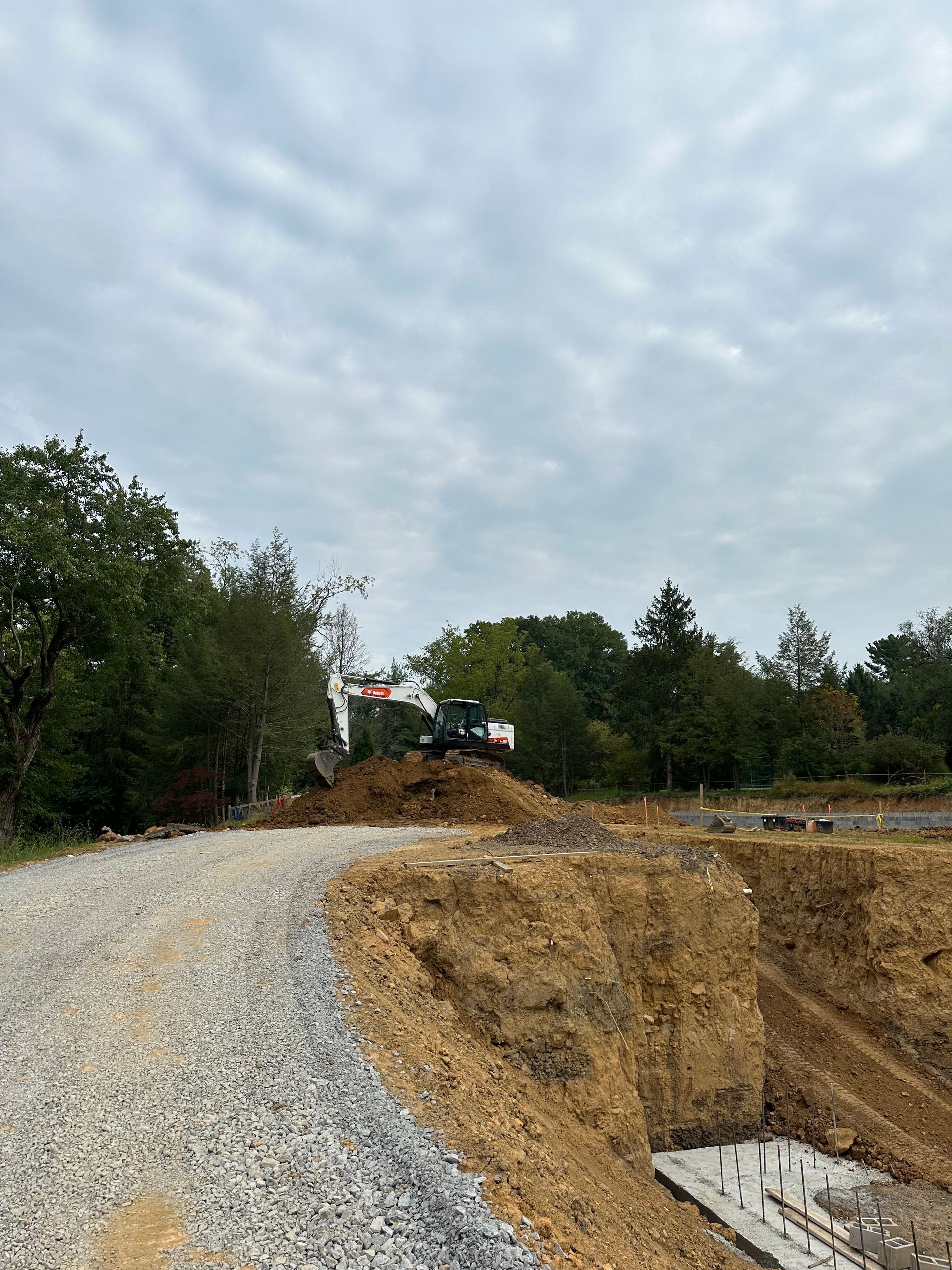 Construction site with excavator on a dirt pile, gravel road, trees, and cloudy sky.