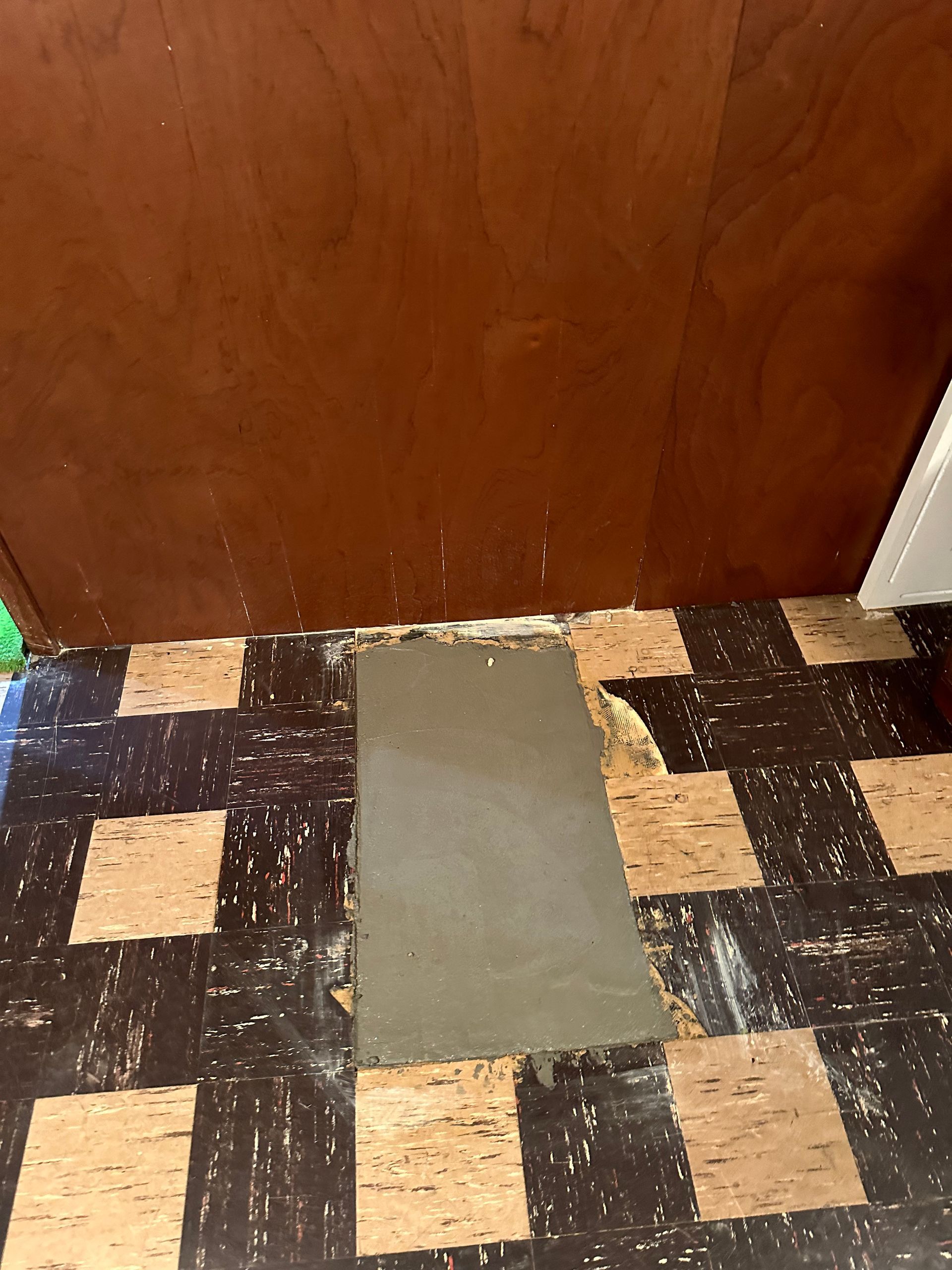 Damaged flooring with a rectangular patch of grey cement, in front of a wood cabinet.