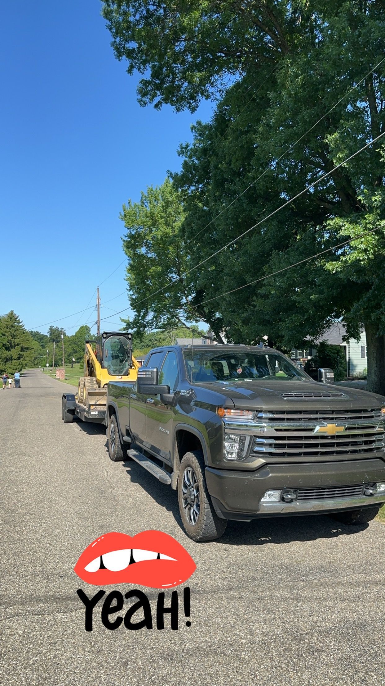 Gray truck with a trailer carrying a small yellow excavator on a gravel road, sunny day.