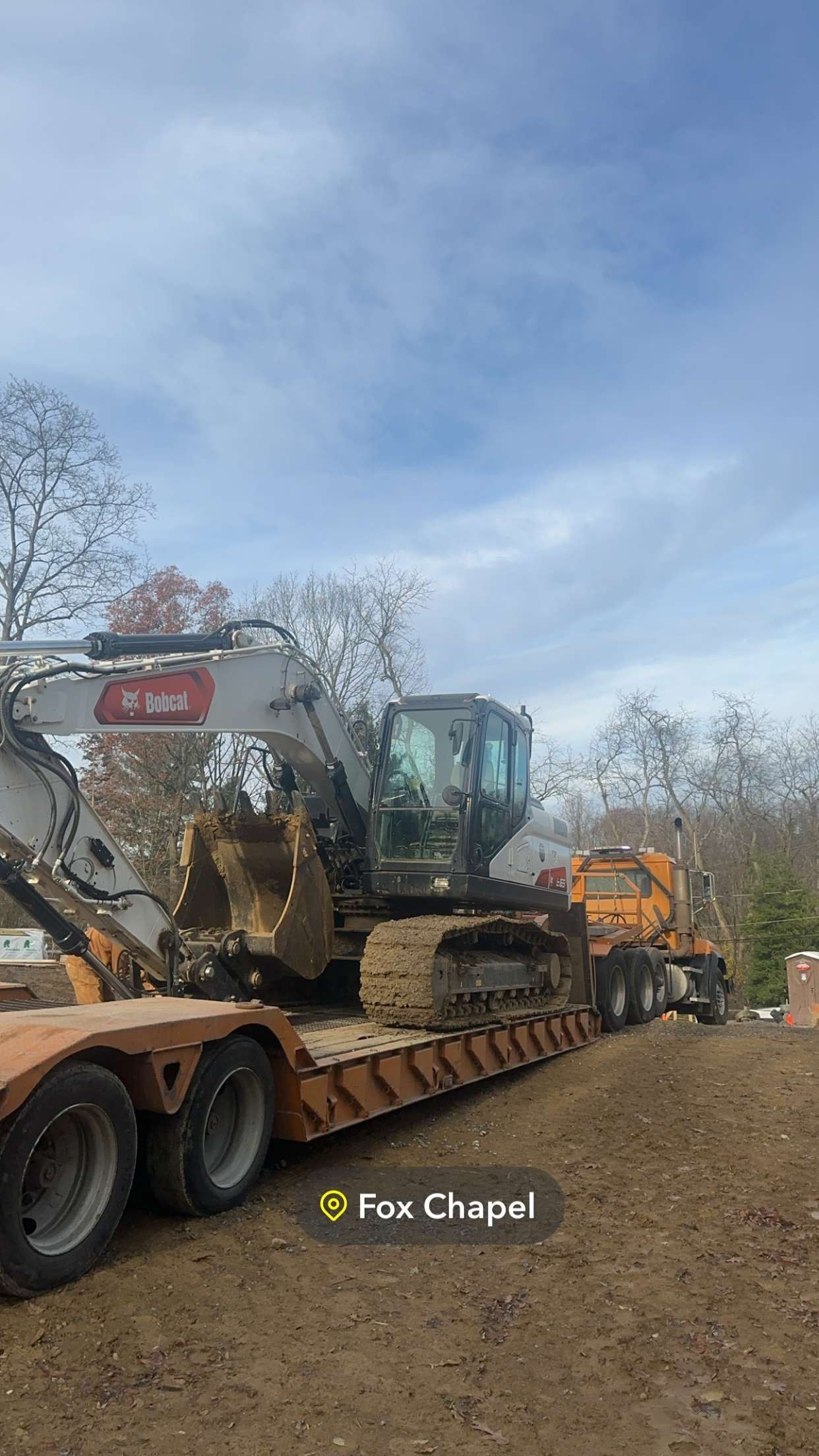 Heavy machinery on a trailer, ready for transport. Tracks, cab, and bucket are visible against a cloudy sky.