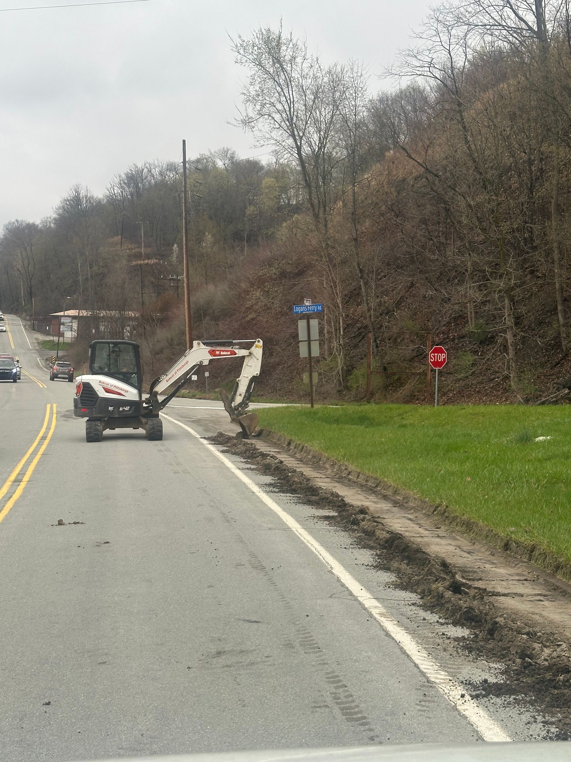 Excavator working on roadside, removing asphalt next to grassy area and road. Hillside in background.