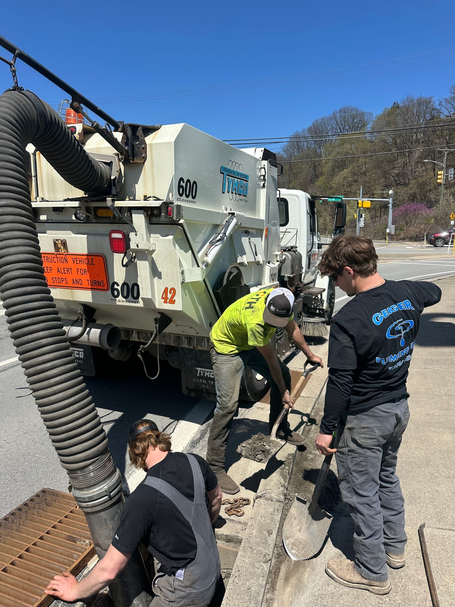 Three workers cleaning a street drain with a vacuum truck. One shovels, one looks on, one tends the hose. Bright sunny day.