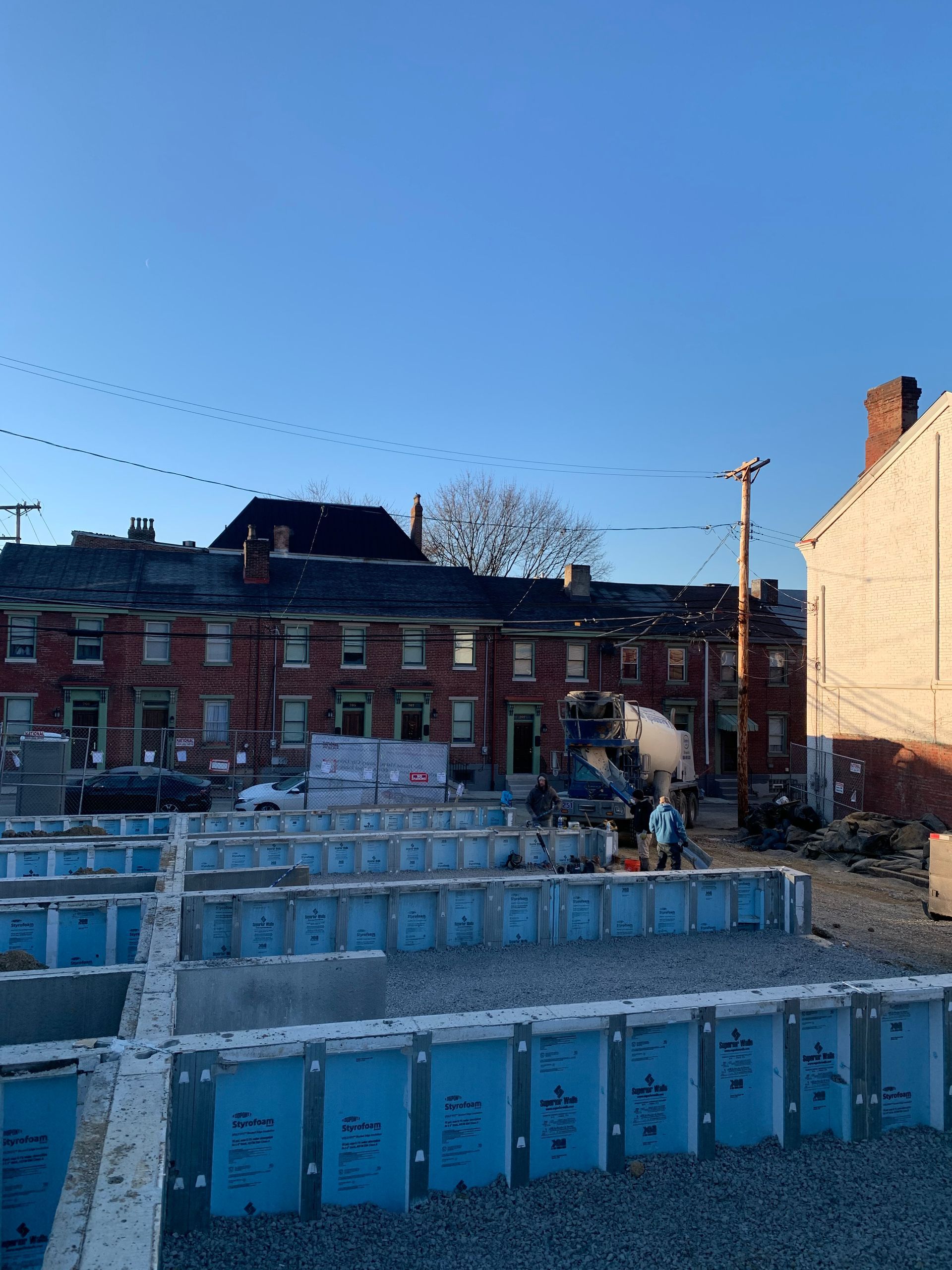 Construction site with foundation forms, brick buildings in background. Clear blue sky.