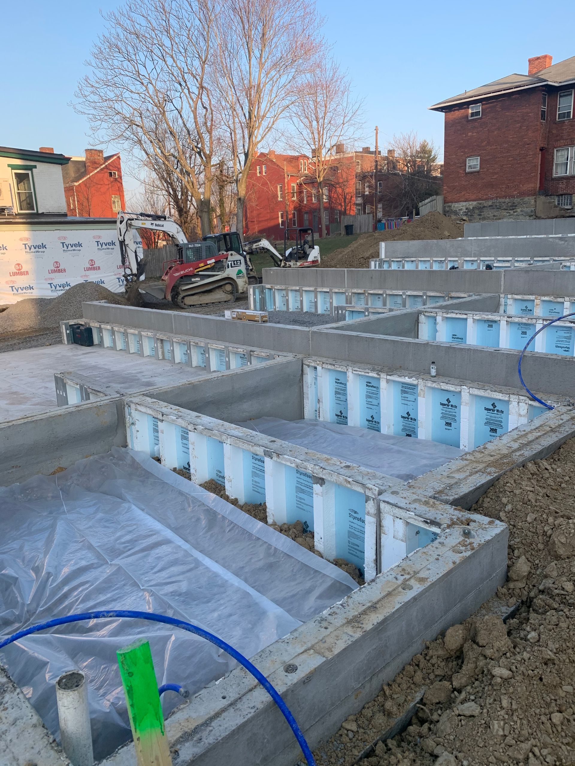 Concrete foundation under construction, with blue insulation panels and heavy machinery in the background.